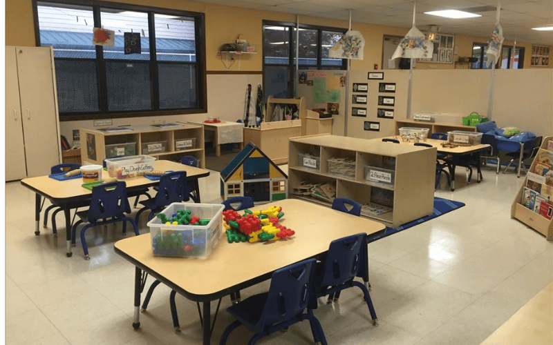 Classroom with small tables, chairs, toy house, and shelving units