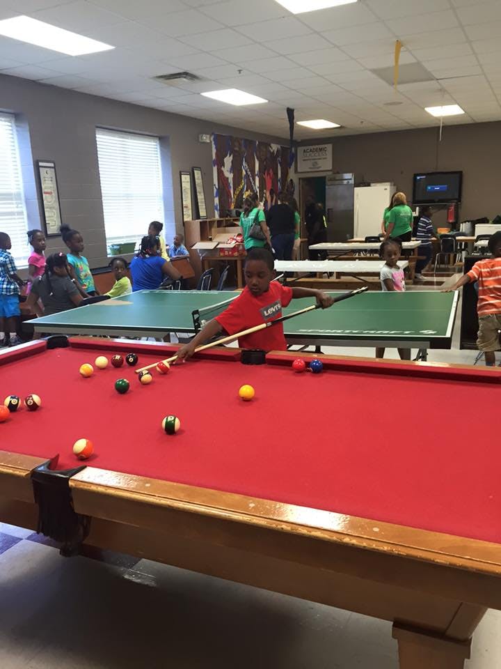 Child playing pool in a room with other people and a ping pong table
