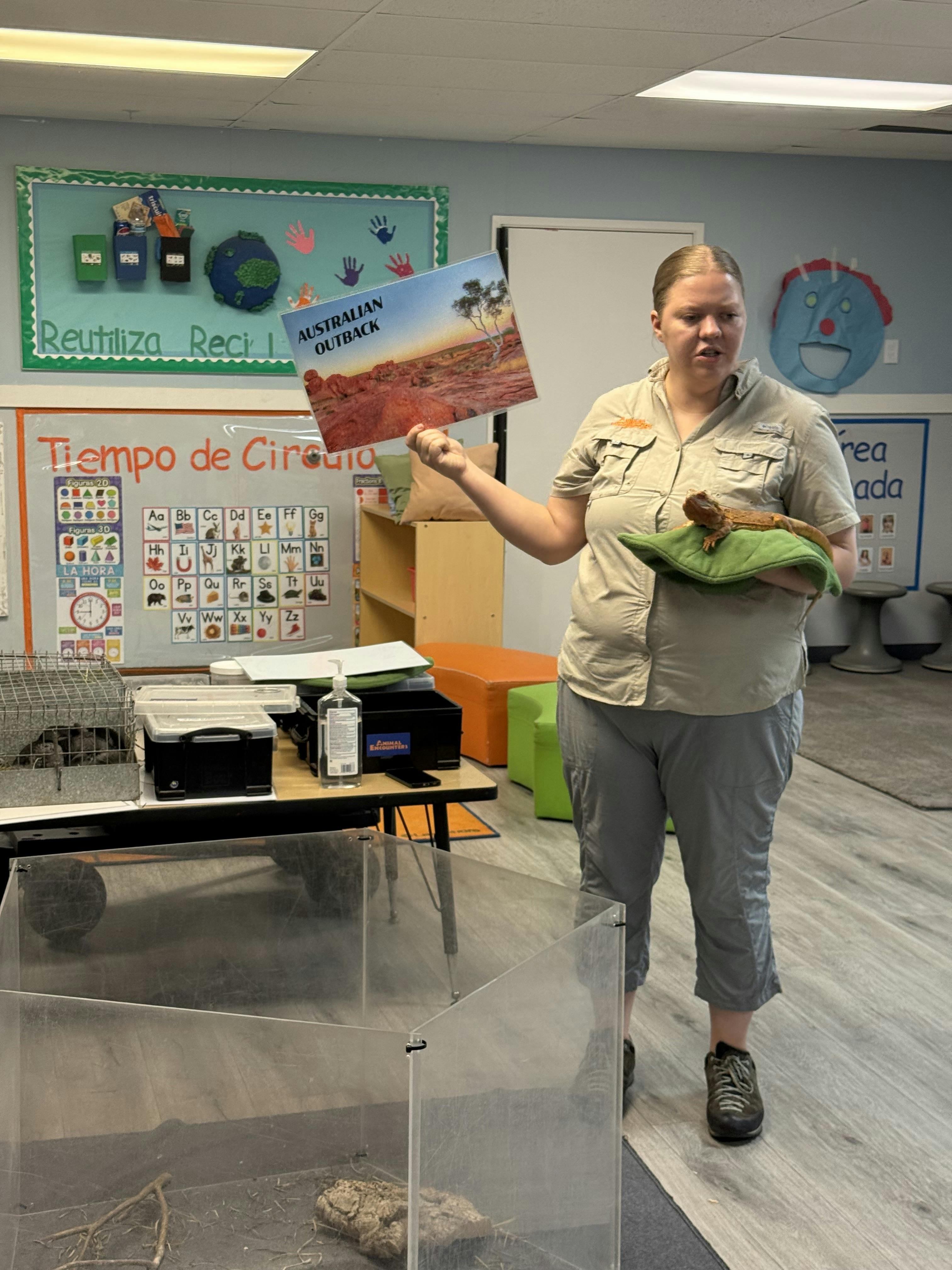 Person holding a lizard and a sign labeled Australian Outback in a classroom