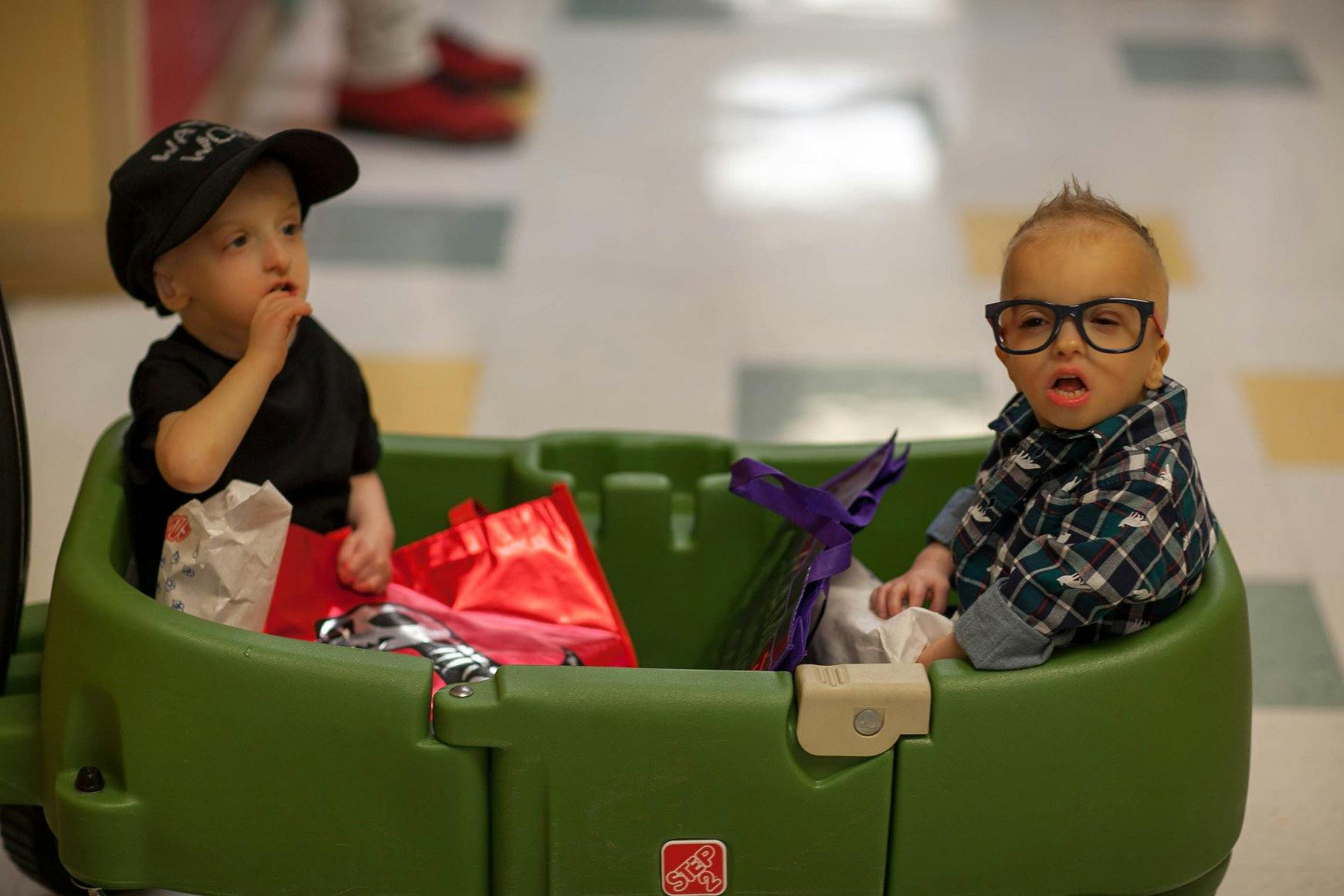 Two children in a green wagon, one wearing a cap, the other wearing glasses