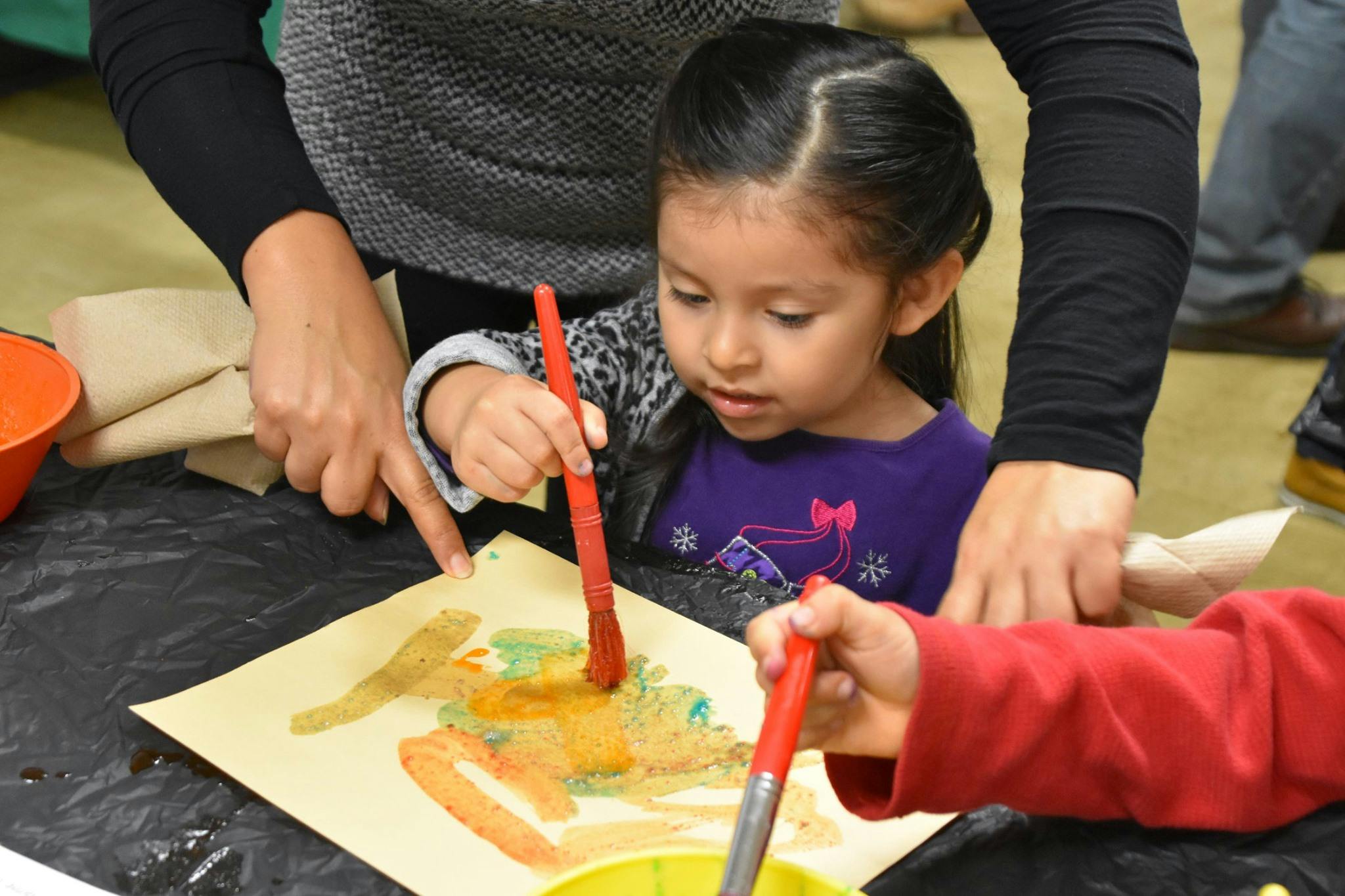 Child painting with a person's hand pointing to the paper