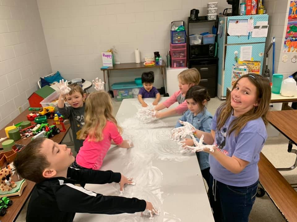 Children playing with white foam on a long table in a classroom