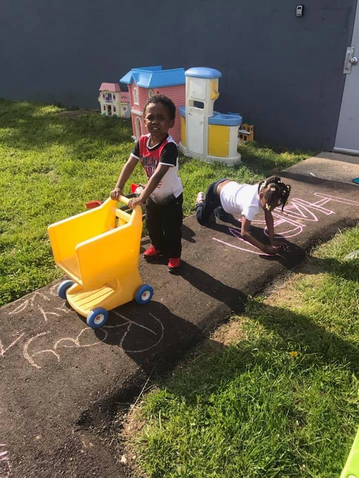 Child pushing a yellow toy shopping cart, another child drawing with chalk