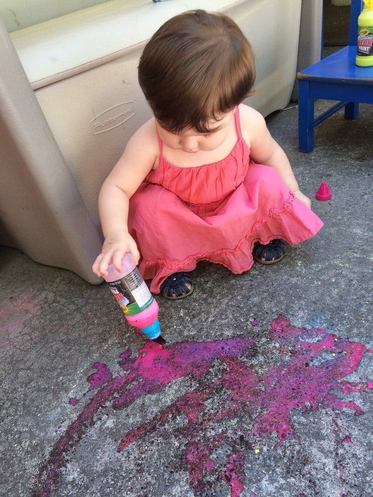 Child in a pink dress drawing on the ground with a colorful marker