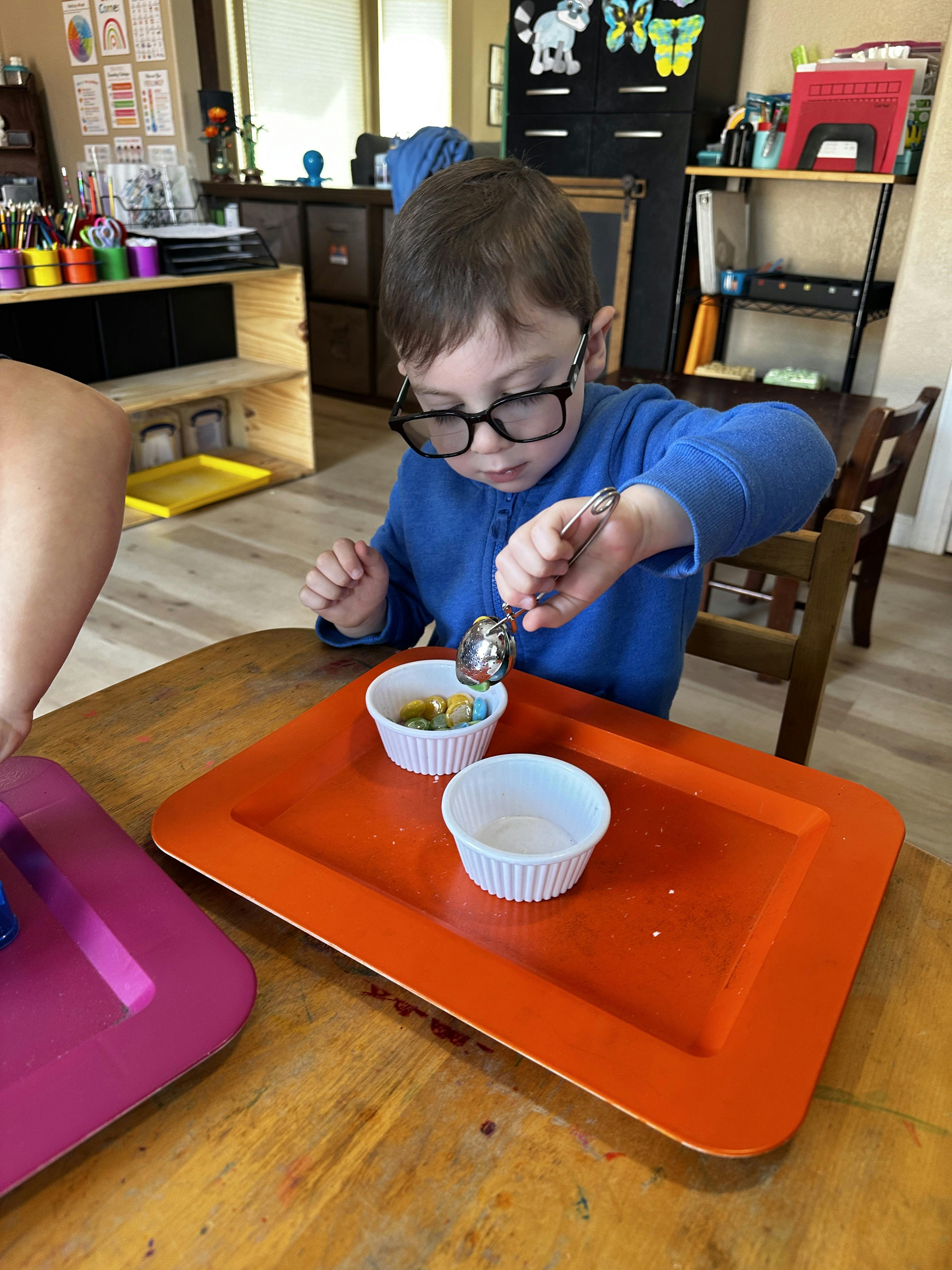 Child wearing glasses transferring small items between containers with a scoop