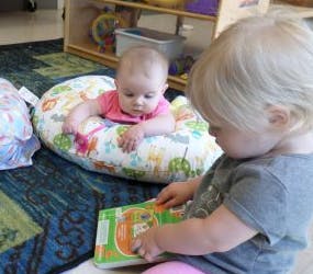 Child reading a book next to a baby in a support pillow