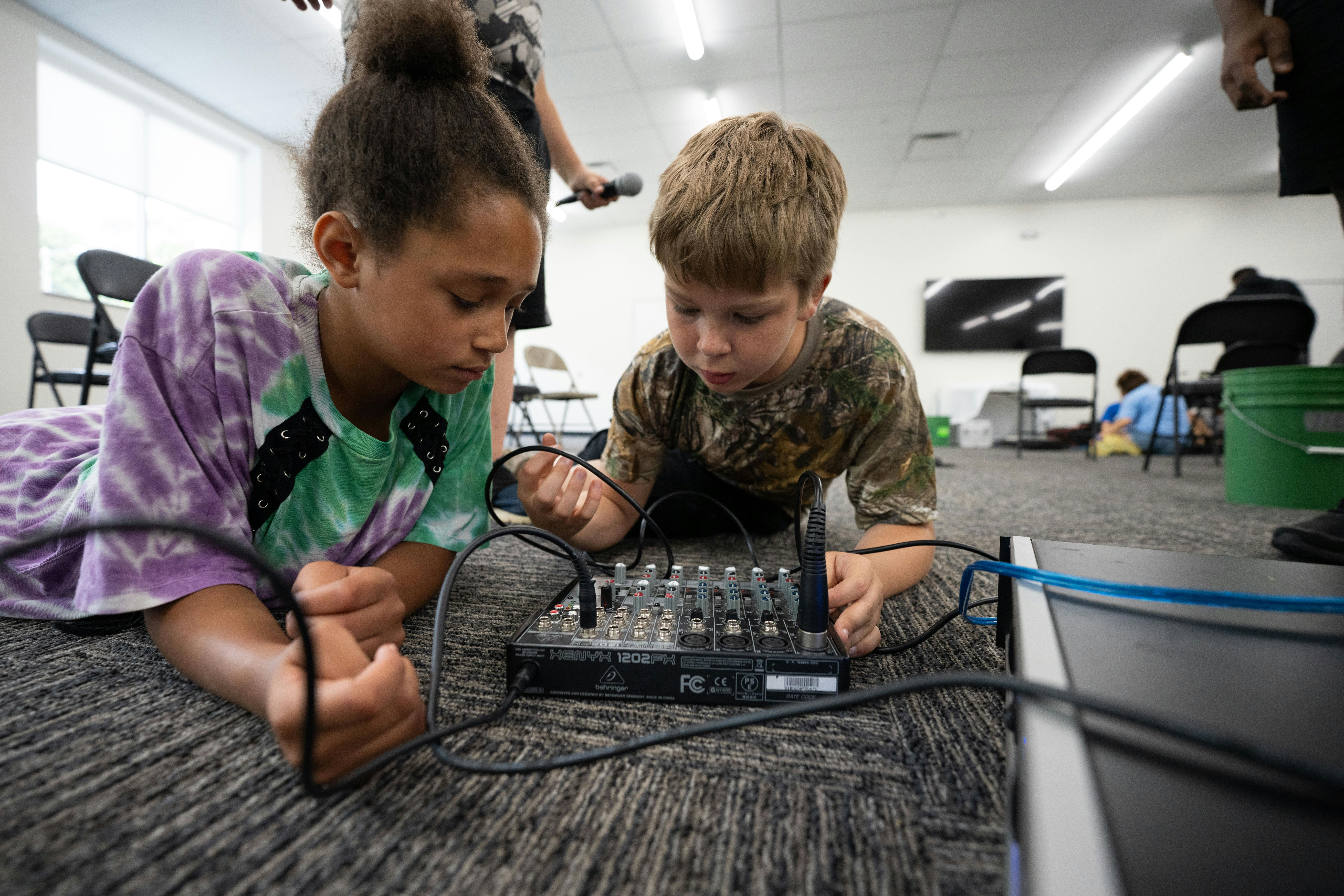 Two children lying on the floor operating an audio mixer with many wires