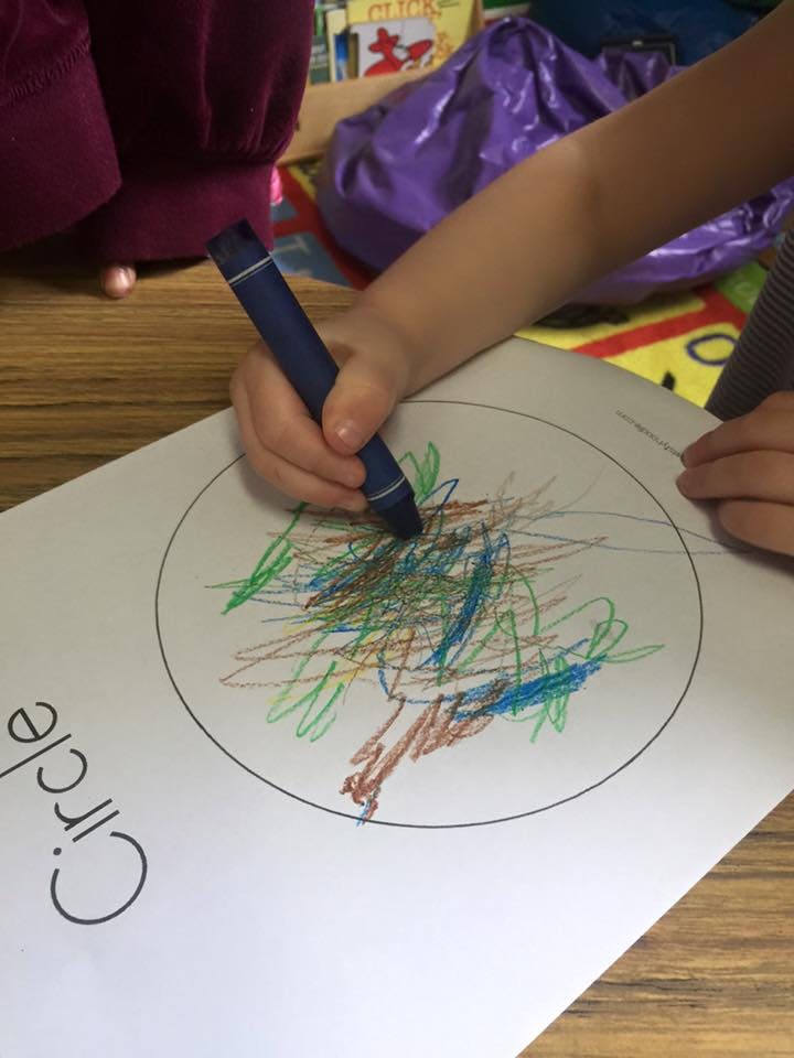 Child's hand holding a crayon and scribbling on paper with a circle outline