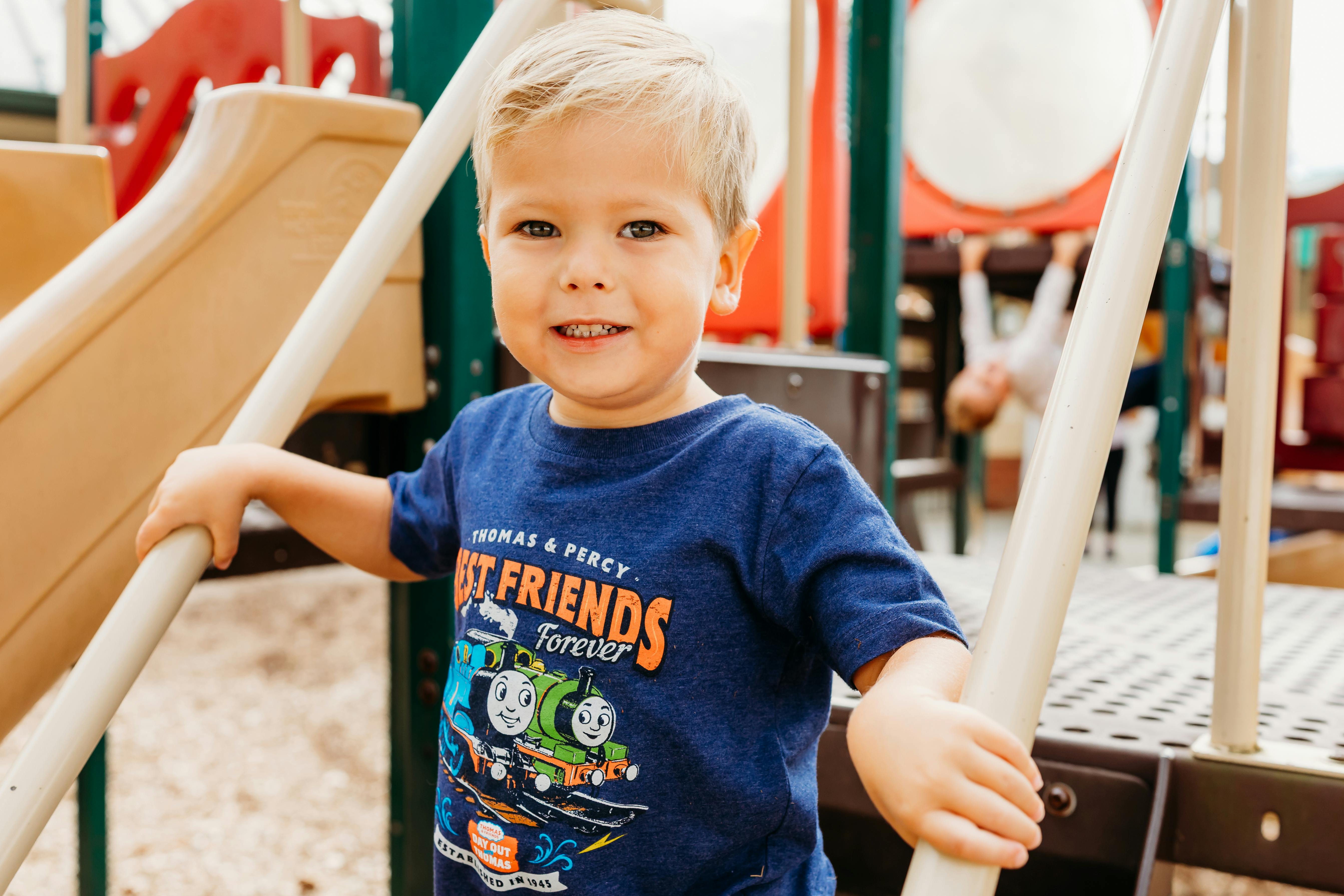 Child in a blue t-shirt with Thomas & Percy holding onto playground bars