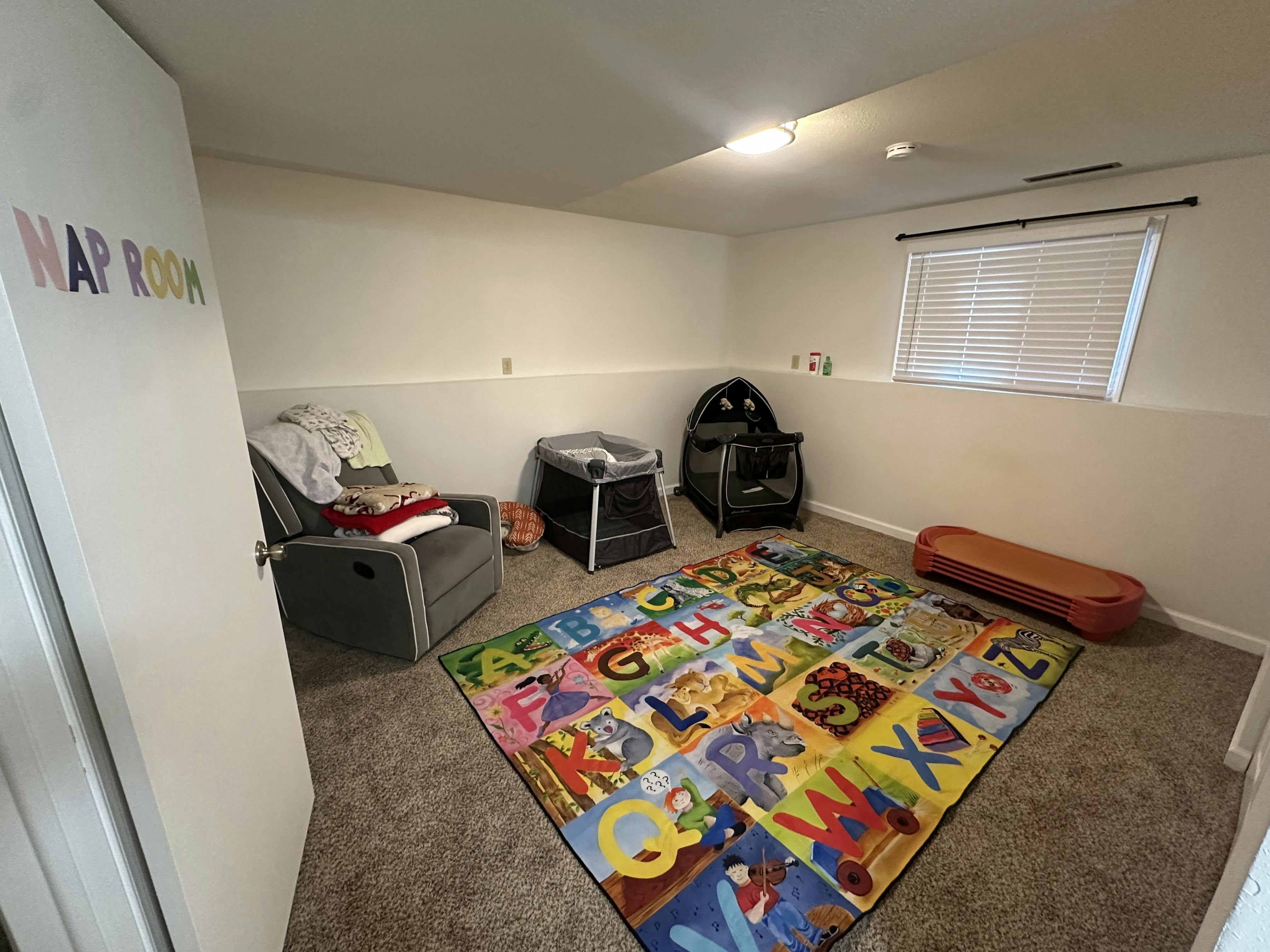 A room with a colorful alphabet rug, a gray recliner, and two playpens