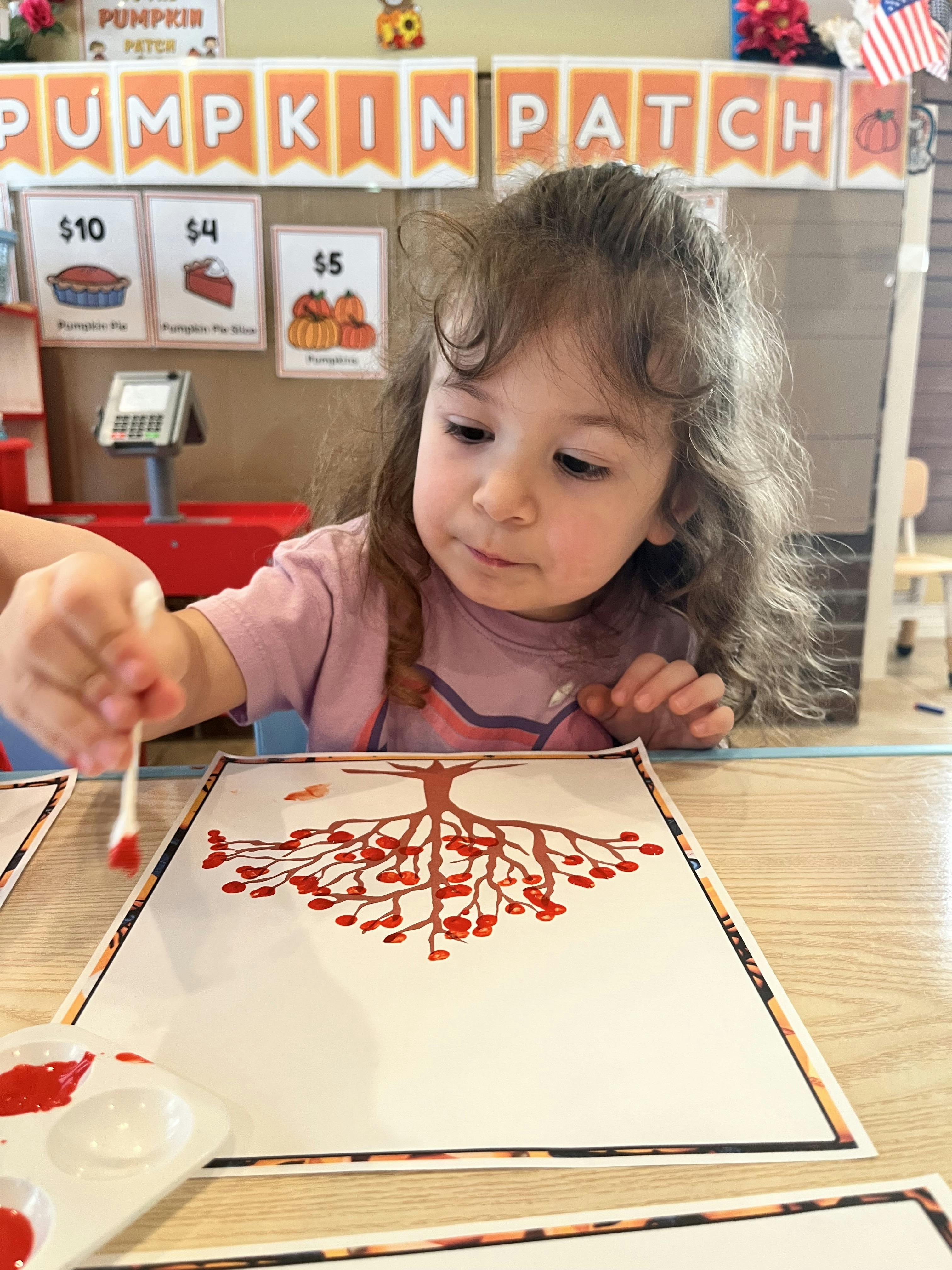 Child painting a tree with red dots on a white paper