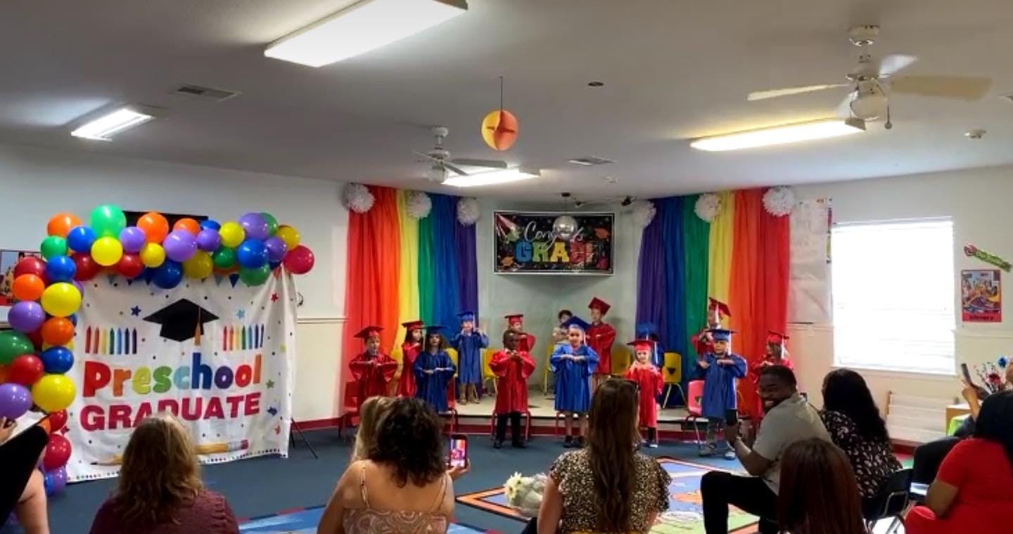 Children in graduation gowns on a stage with a "Preschool Graduate" banner