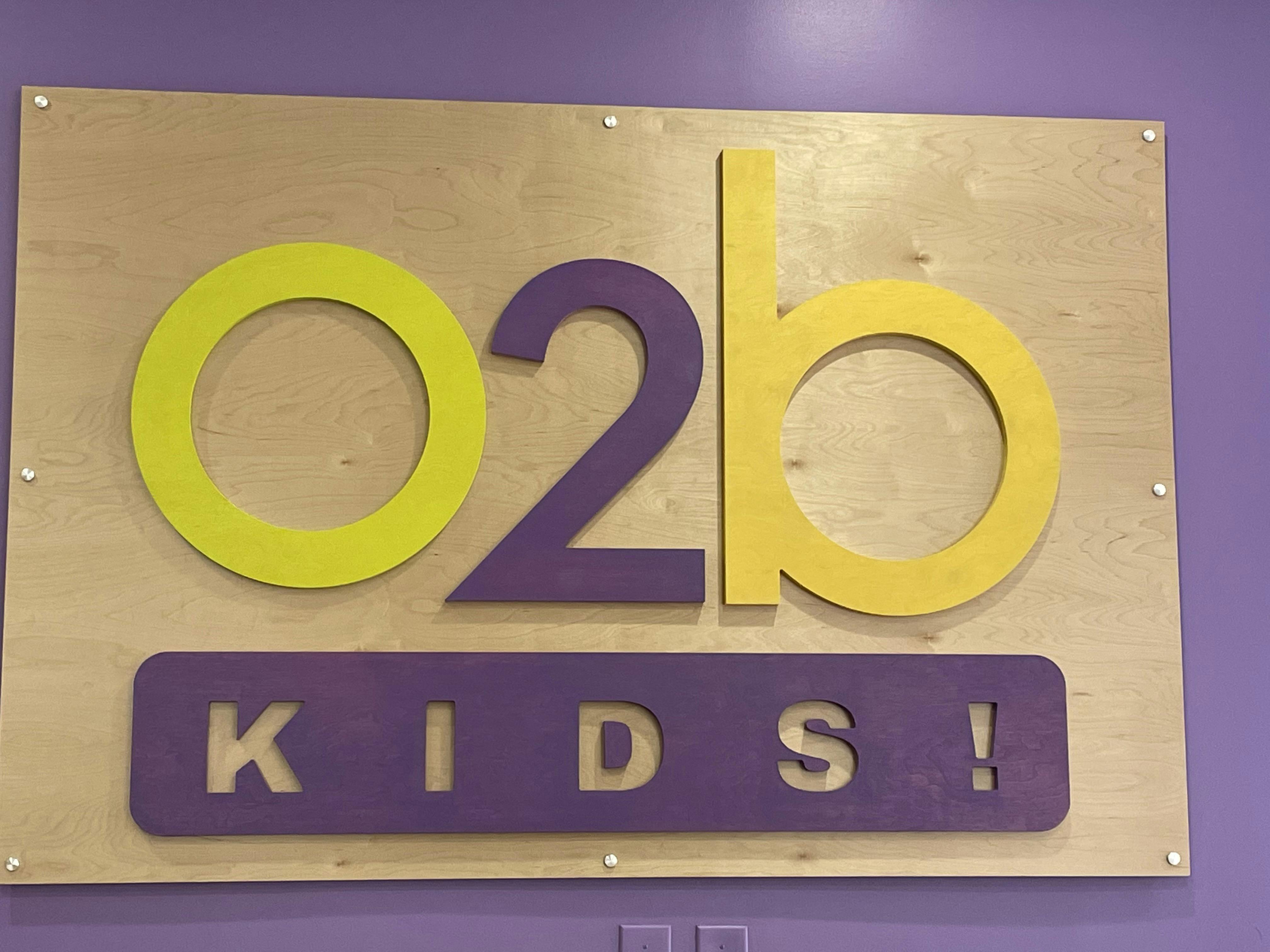 Sign with \"O2b Kids!\" in yellow and purple letters on a wooden board