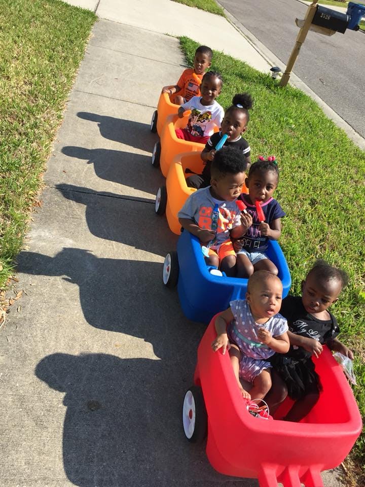 Multiple children sitting in a line of connected wagons on a sidewalk