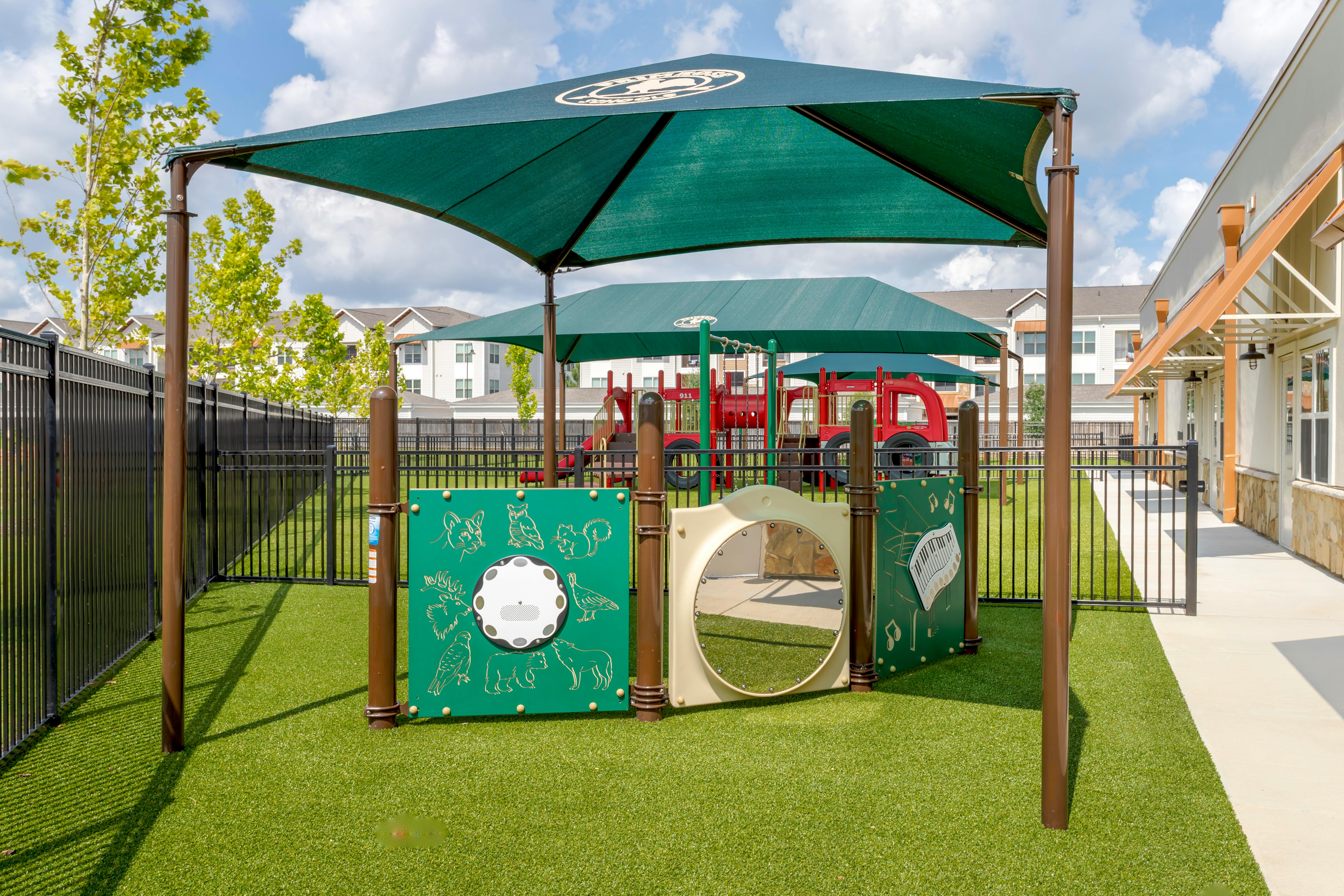 Playground with green turf, shaded play structures, and a black fence