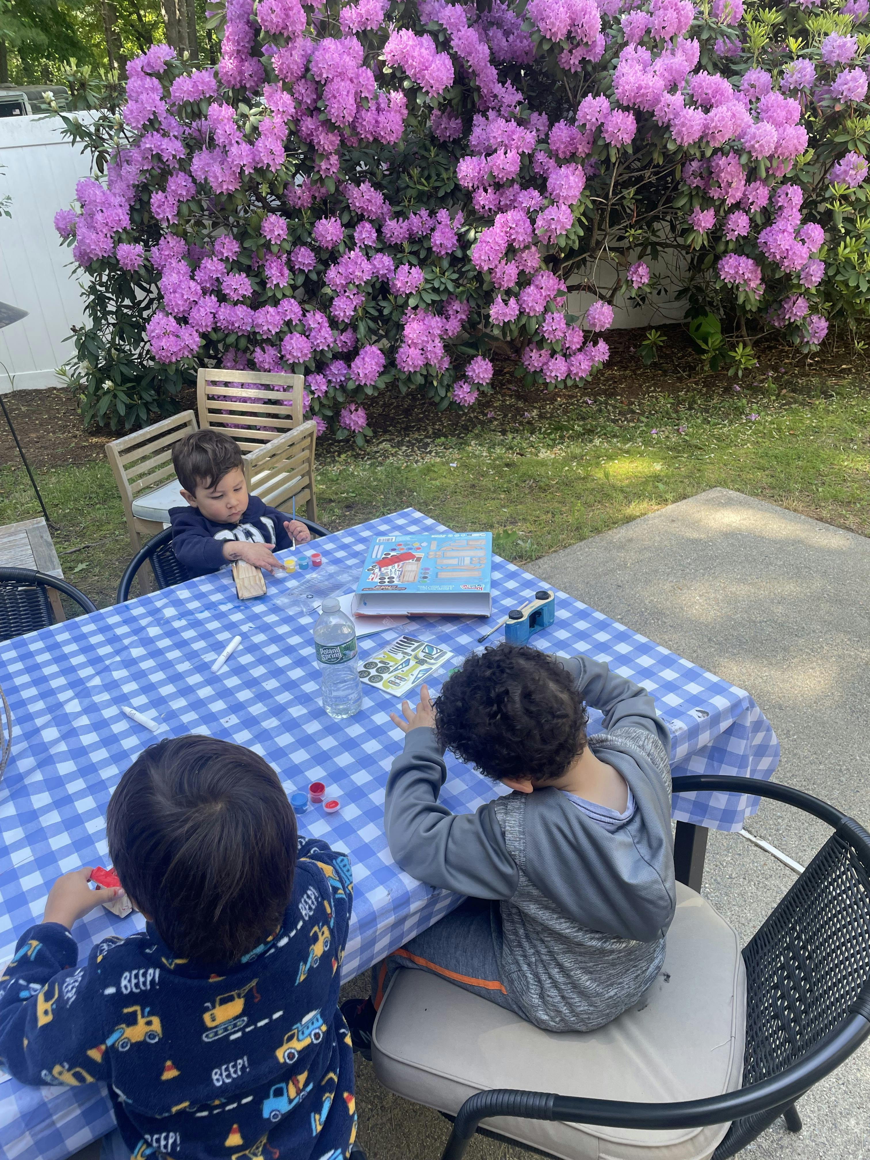 Three children painting at an outdoor table with a large bush of purple flowers