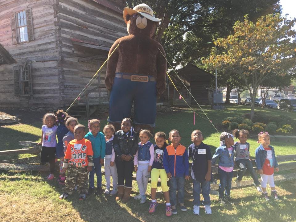 Children standing in front of a large Smokey Bear statue and a log cabin