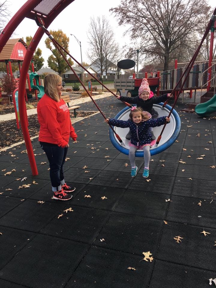 Person standing next to two children on a swing in a playground