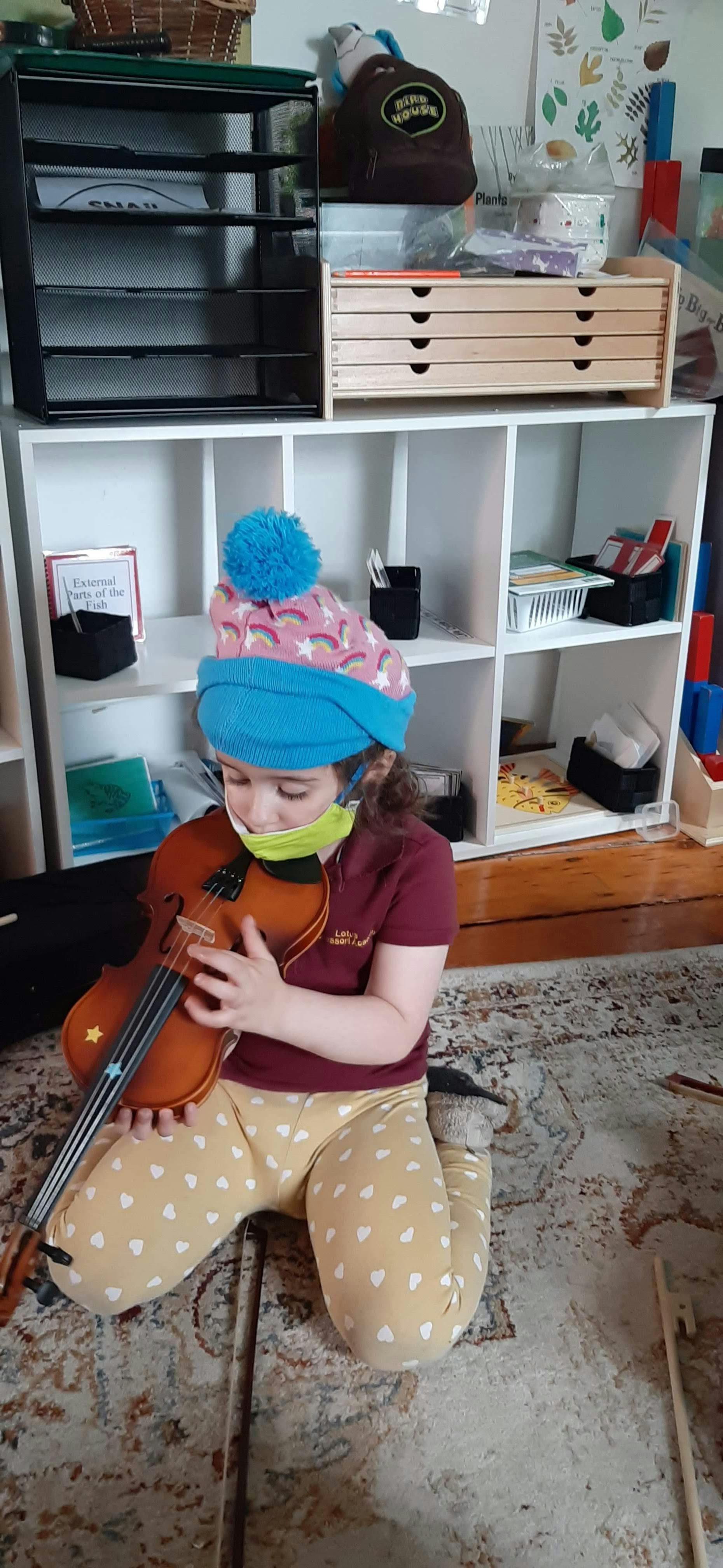 Child in a knit hat and mask playing a violin while sitting on the floor