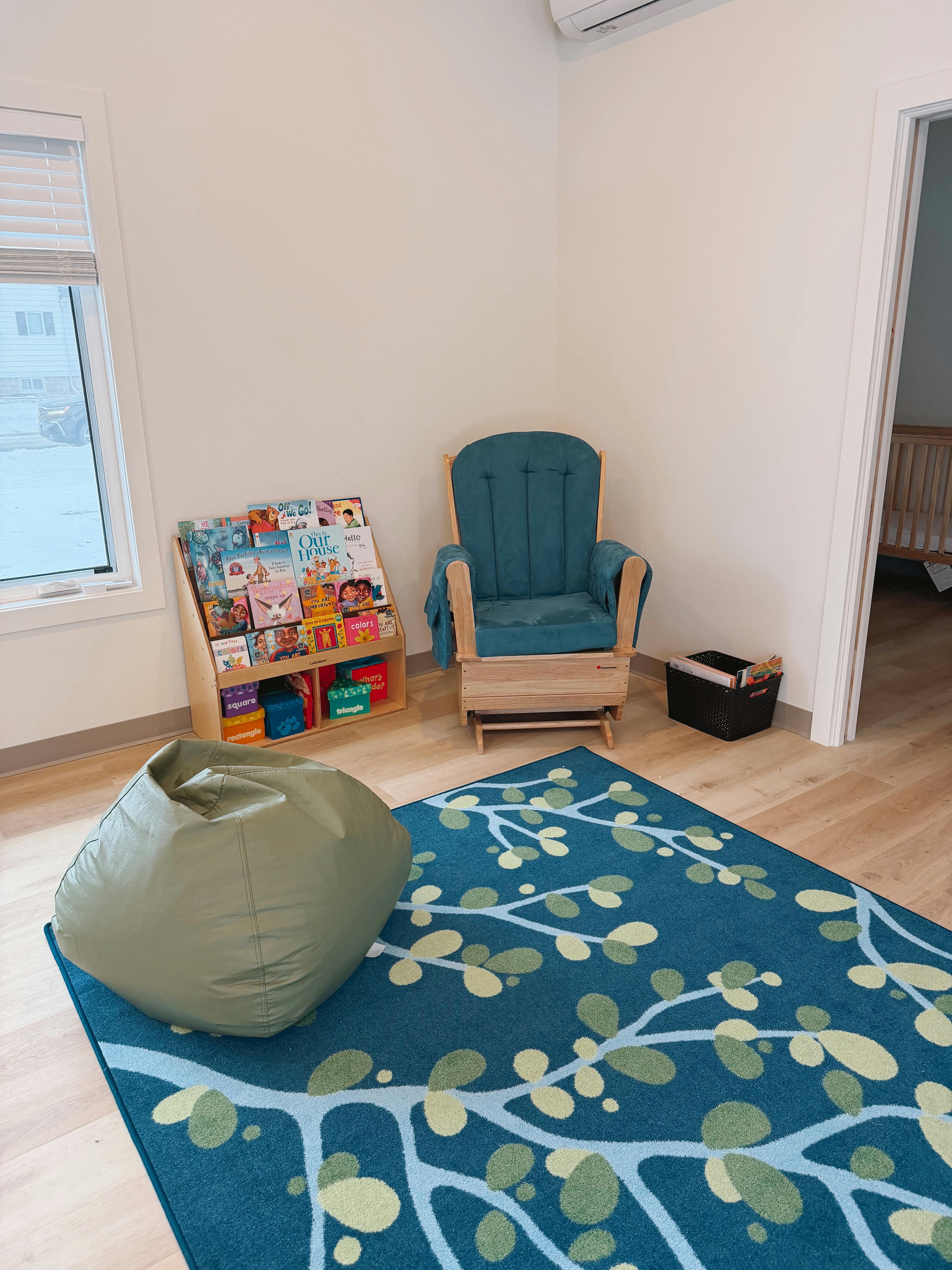 A children's reading nook with a teal rug, beanbag, rocking chair, and bookcase