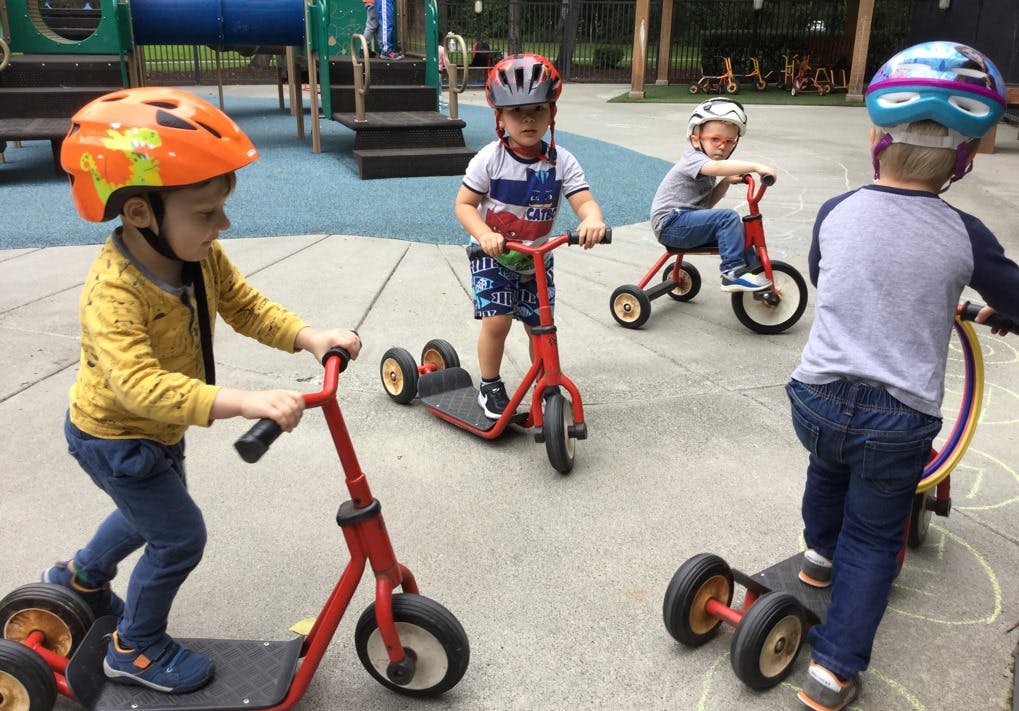 Children wearing helmets riding scooters and tricycles on a playground