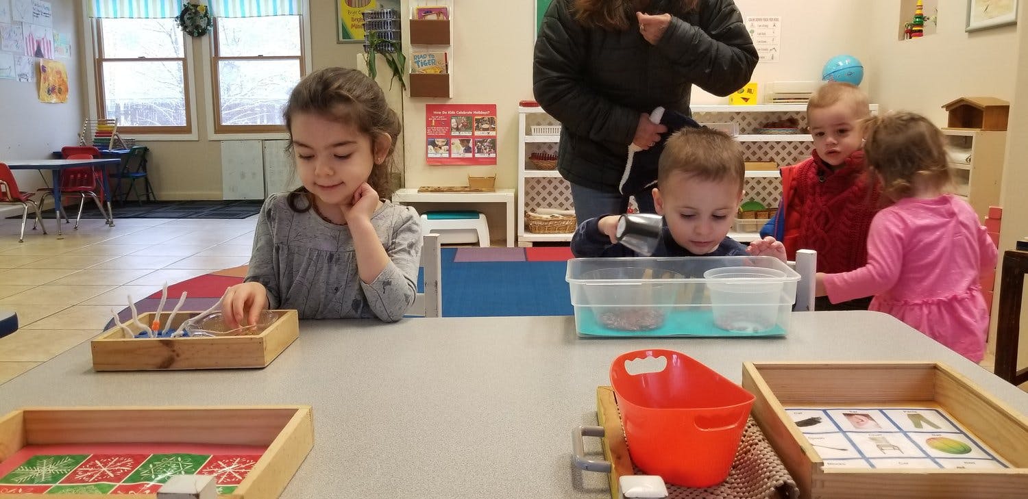 Children at a table in a classroom, one child playing with sticks in a box