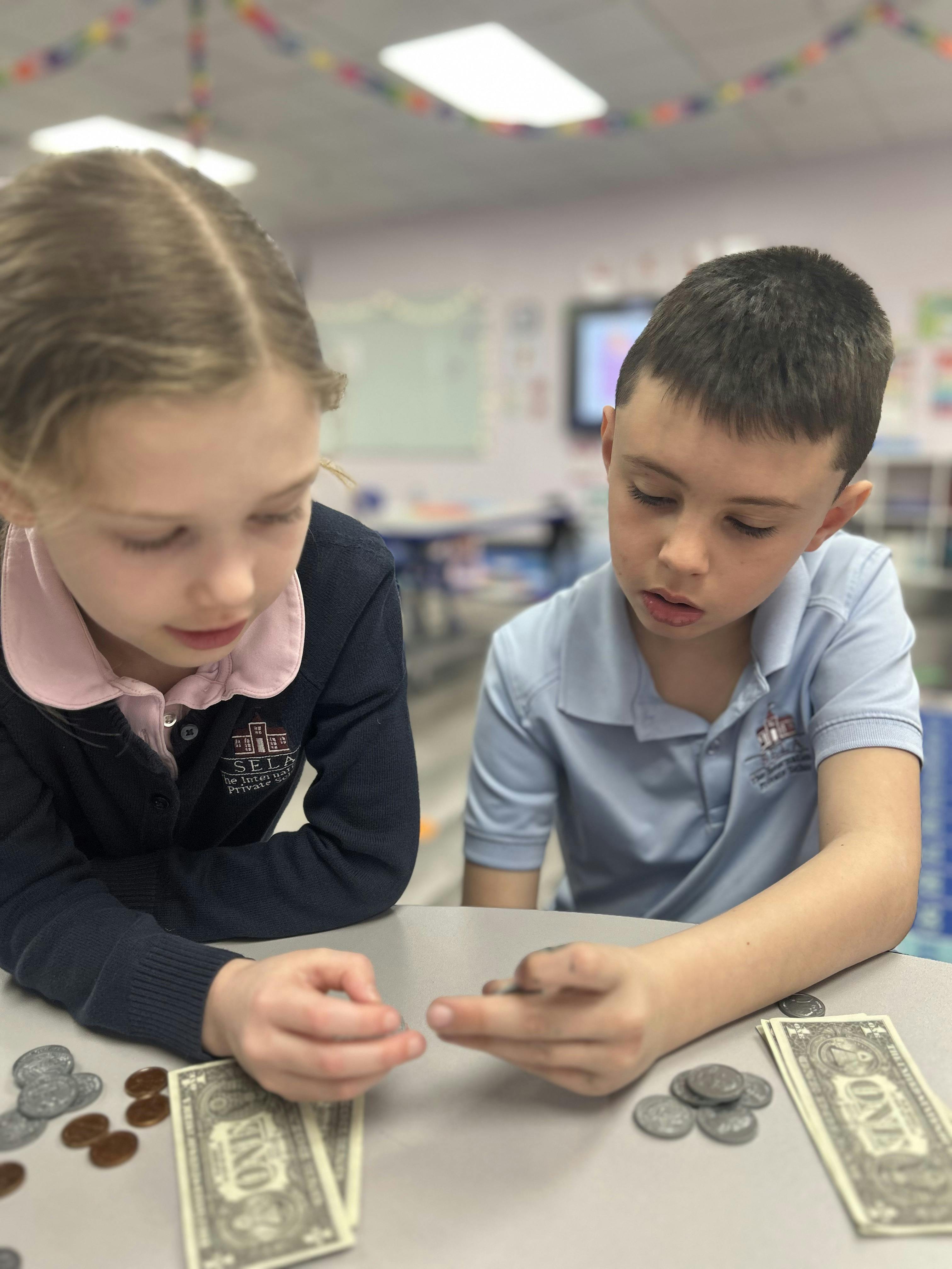 Two children counting money on a table in a classroom