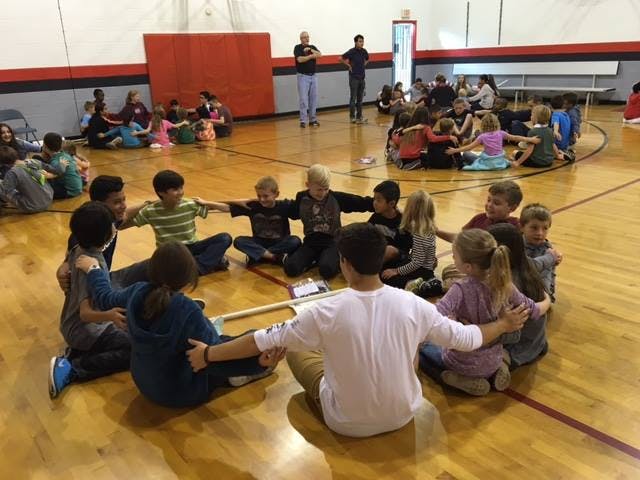 Children and adults sitting in a circle holding hands on a gym floor