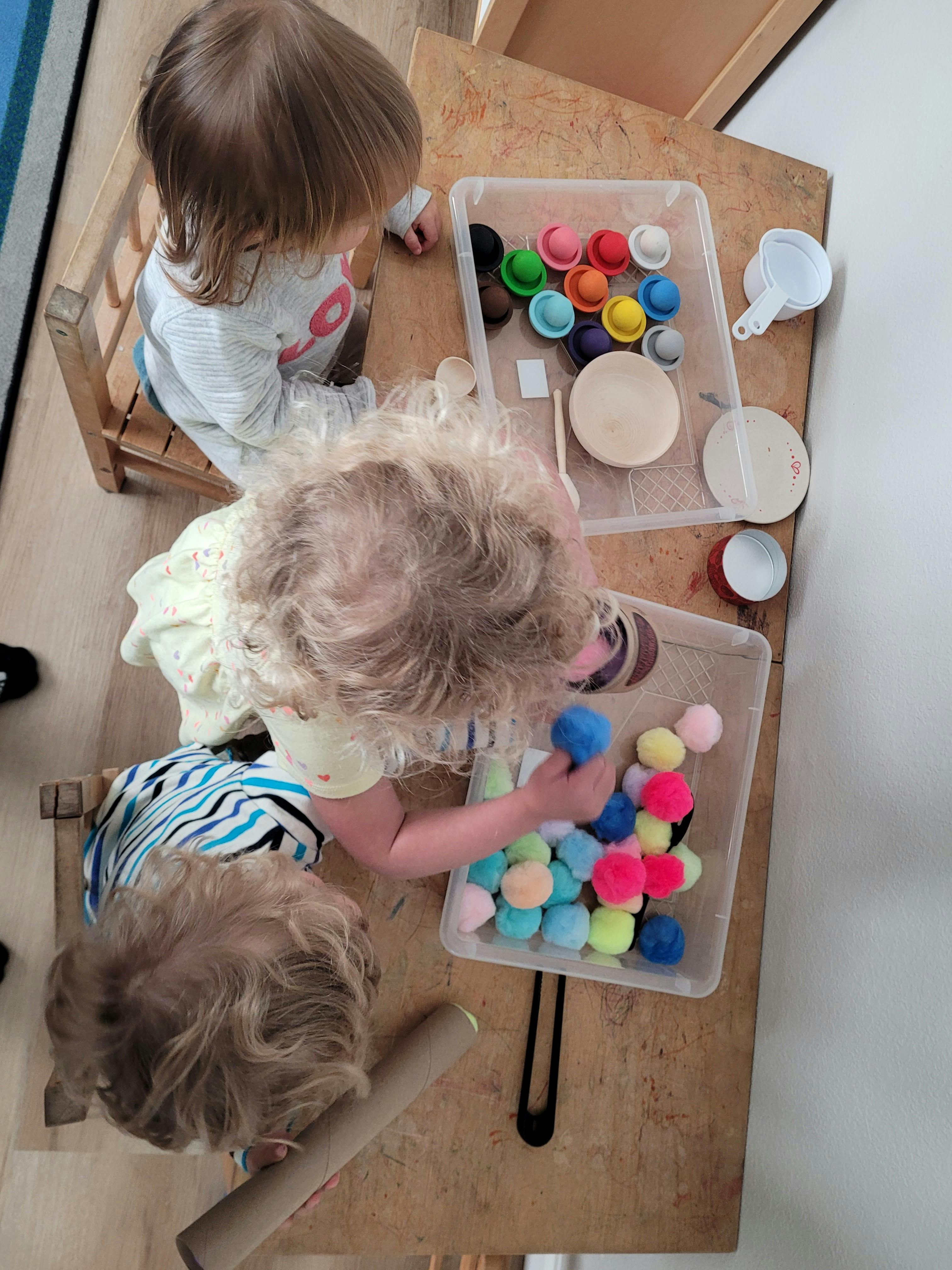 Three children playing with colorful pom-poms and small cups on a table