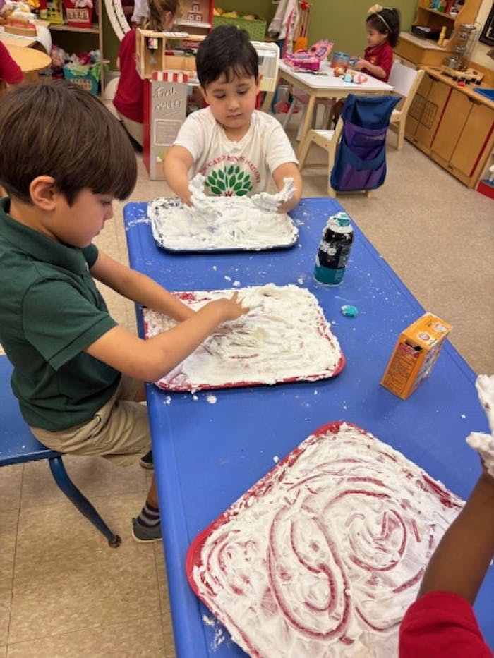 Children playing with shaving cream on trays at a blue table in a classroom