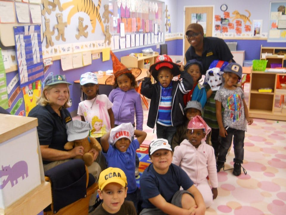 Two adults and multiple children in a classroom, many wearing hats