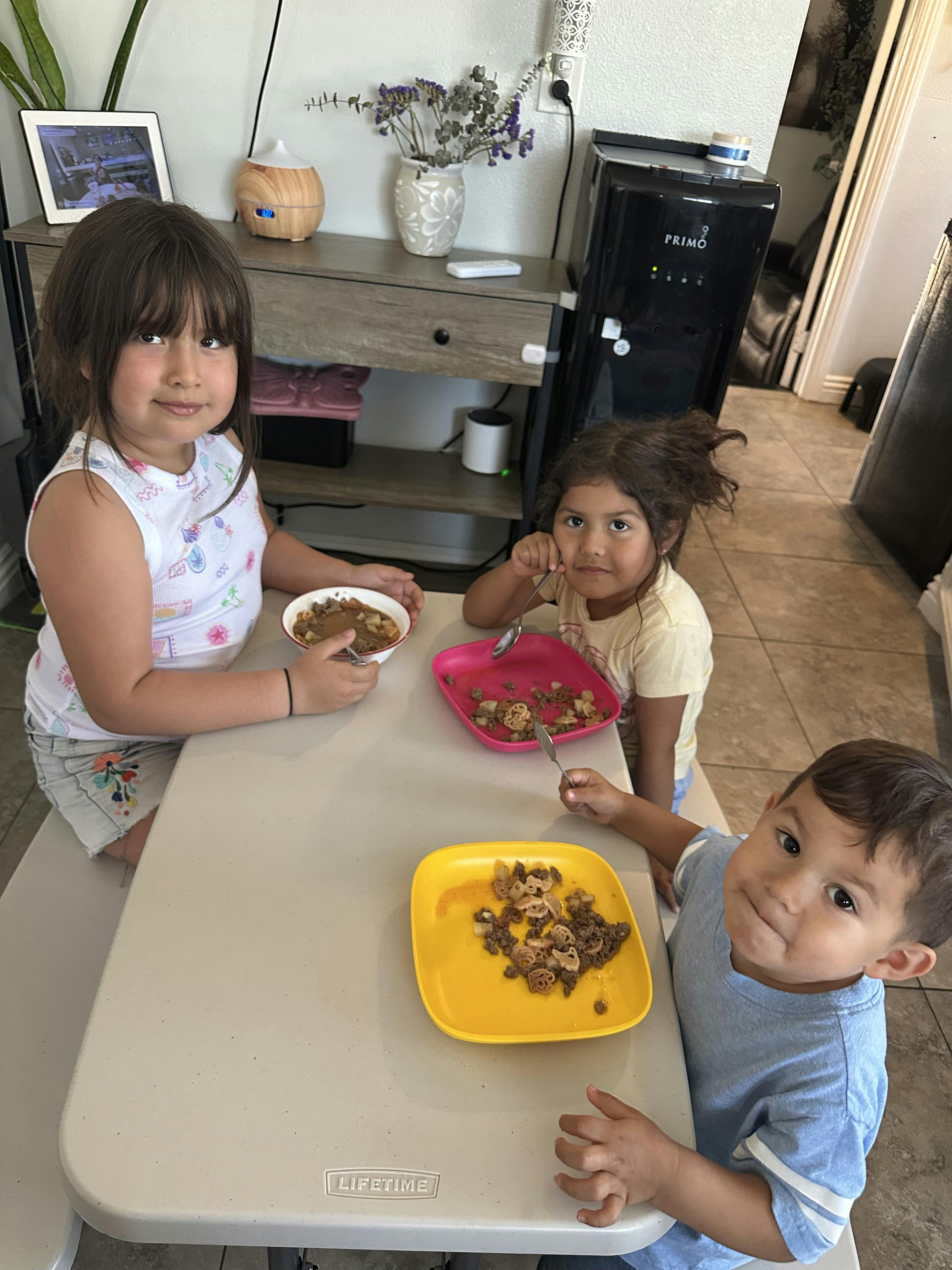 Three children sitting at a table eating meals from colorful plates and bowls