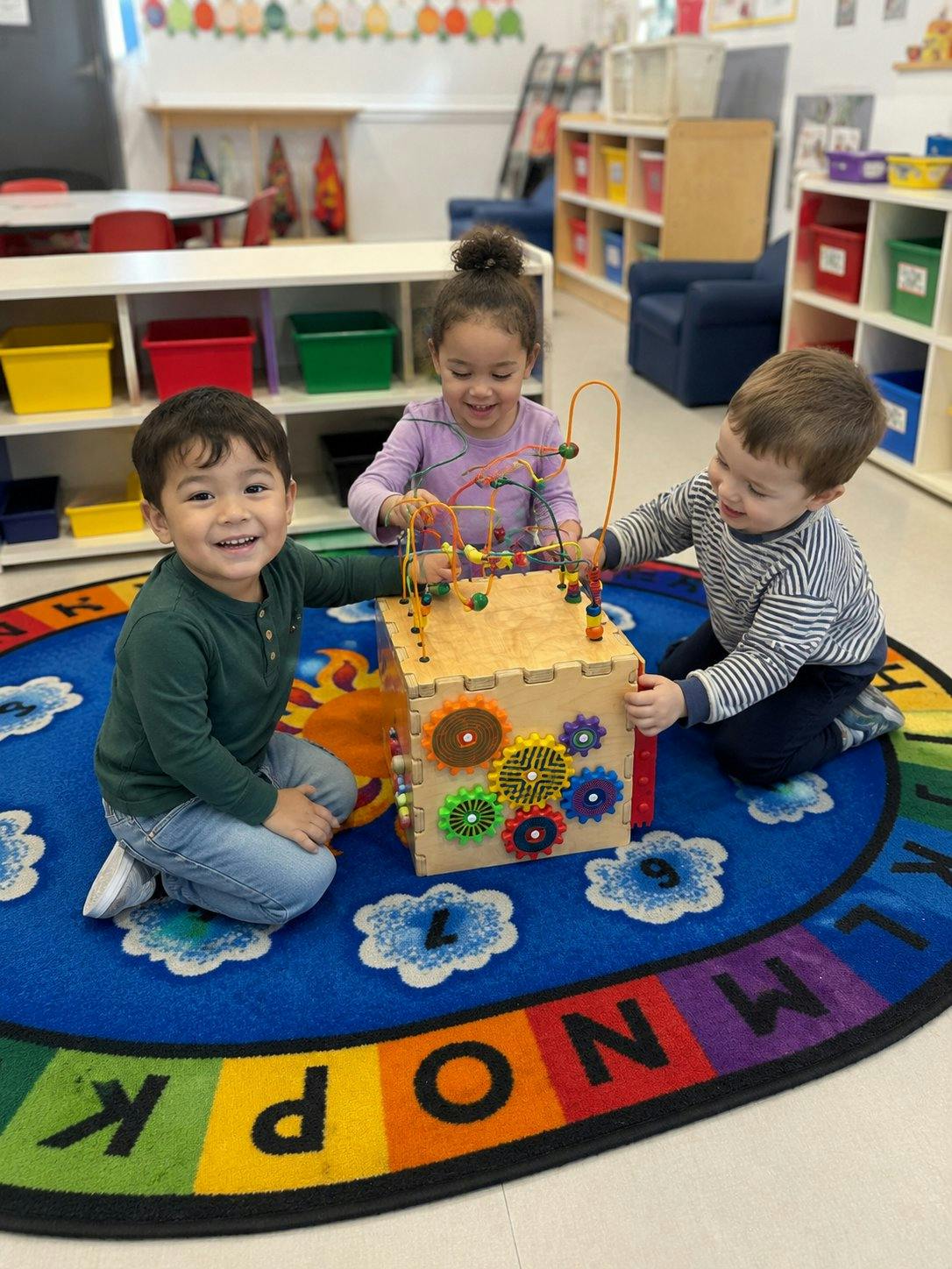 Three young children playing together with an activity cube on a classroom rug