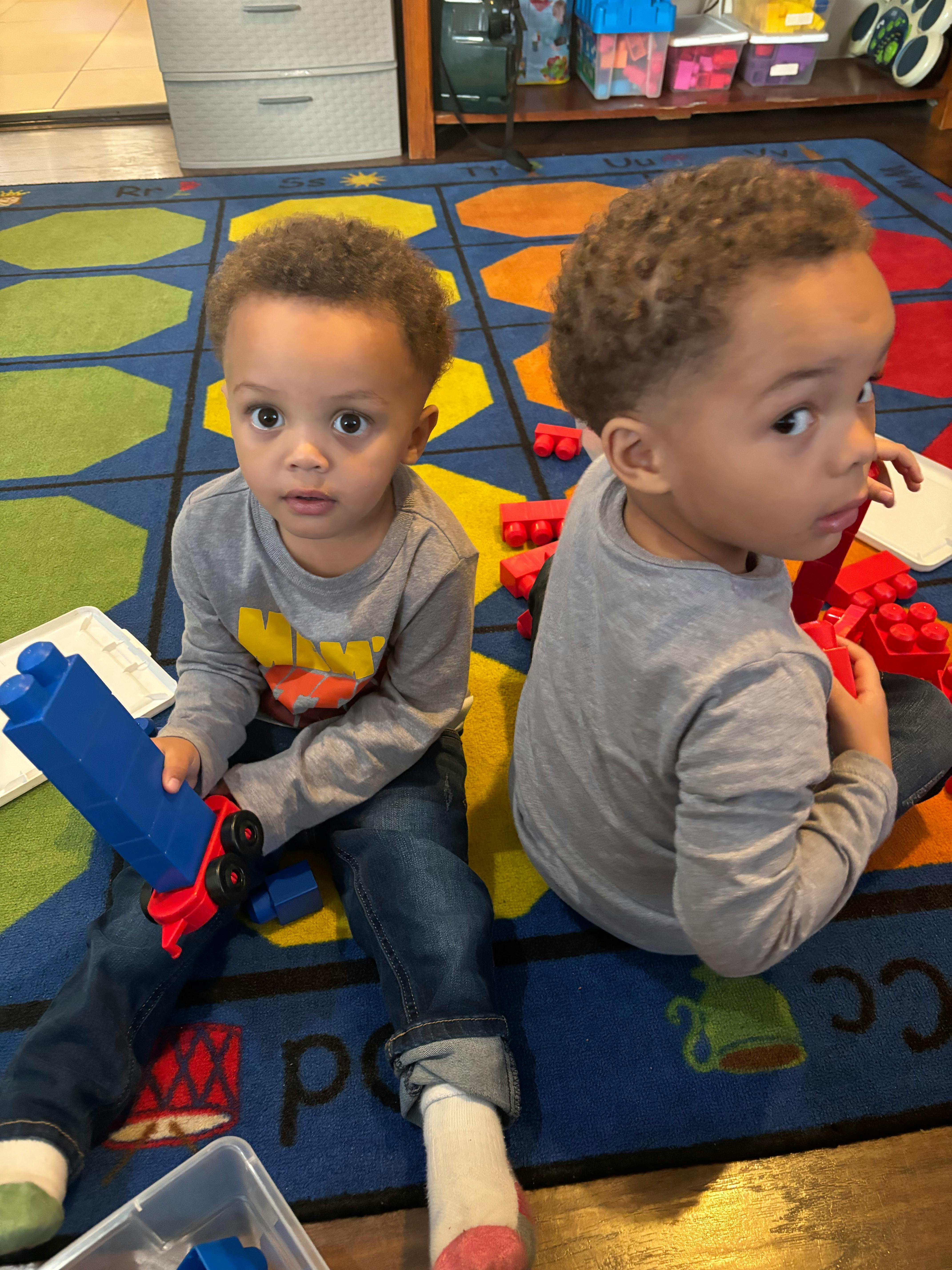 Two children sitting on a colorful rug, one holding a blue toy