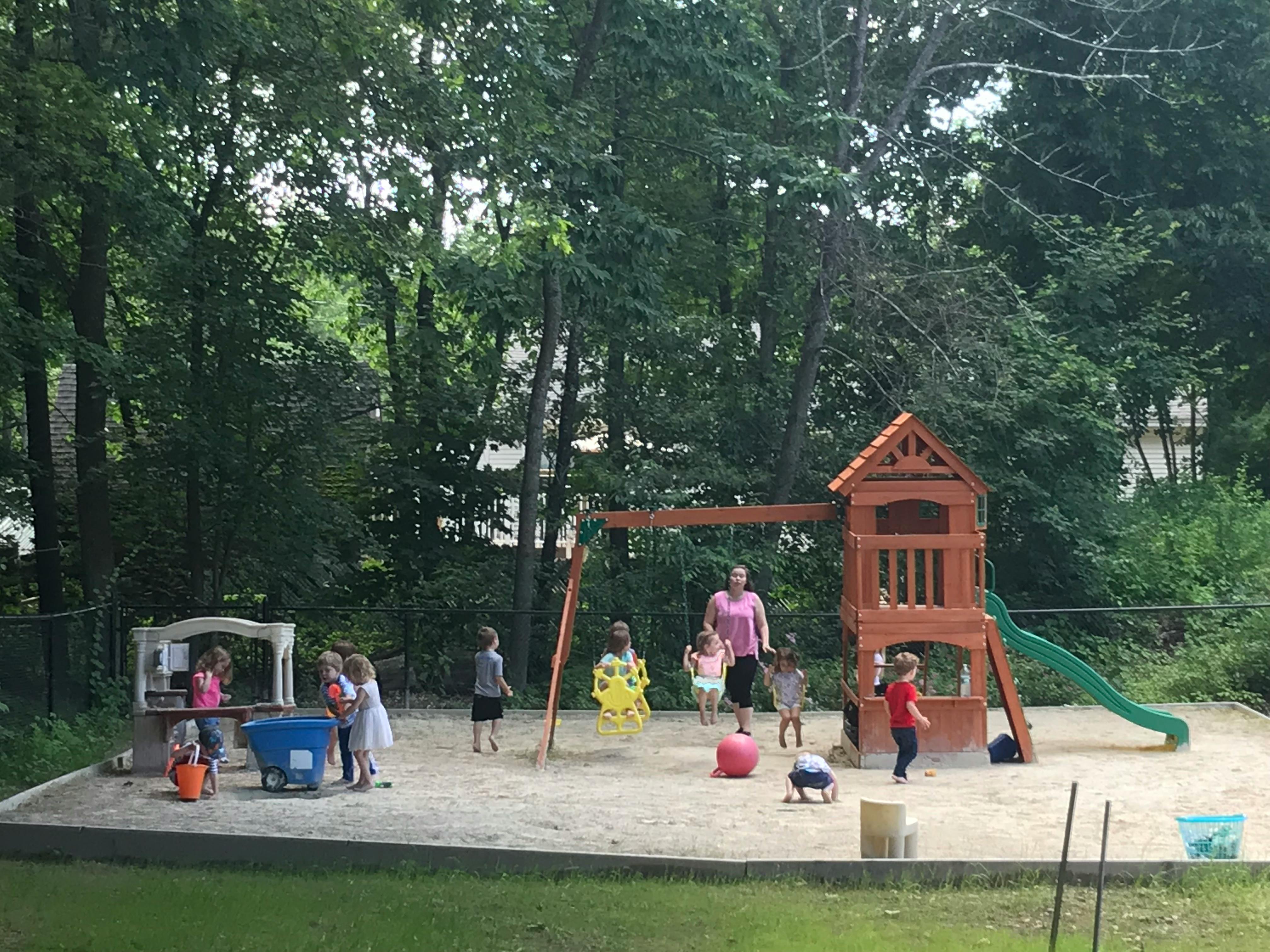 Children and an adult playing in a sandbox with a swing set and playhouse