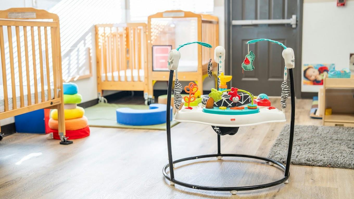 Baby jumper with hanging toys in a daycare room with cribs
