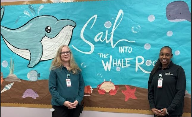 Two people standing in front of a mural with a dolphin and text "Sail into the Whale Ro"