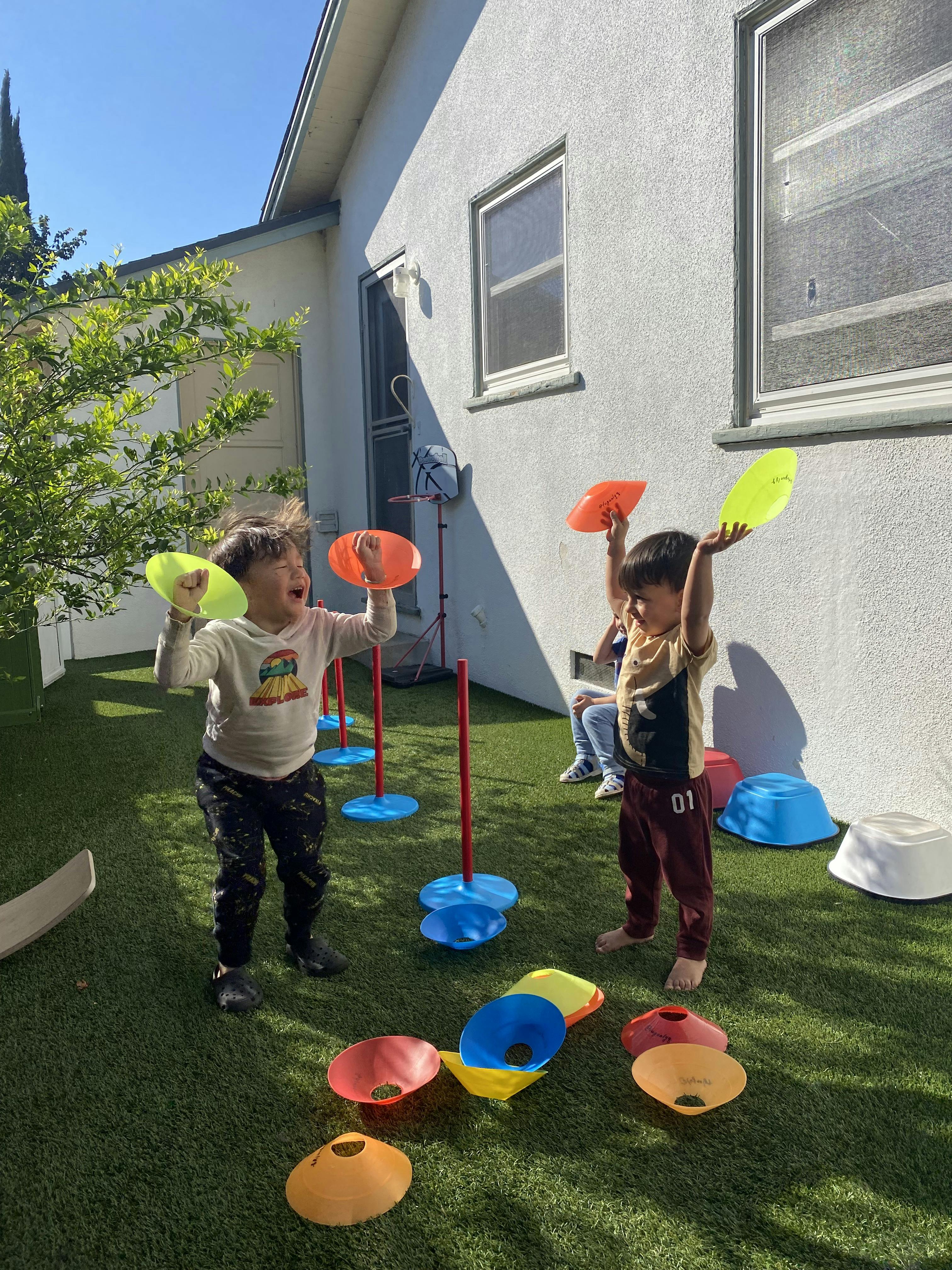 Two children playing with colorful plastic discs on artificial grass in a backyard