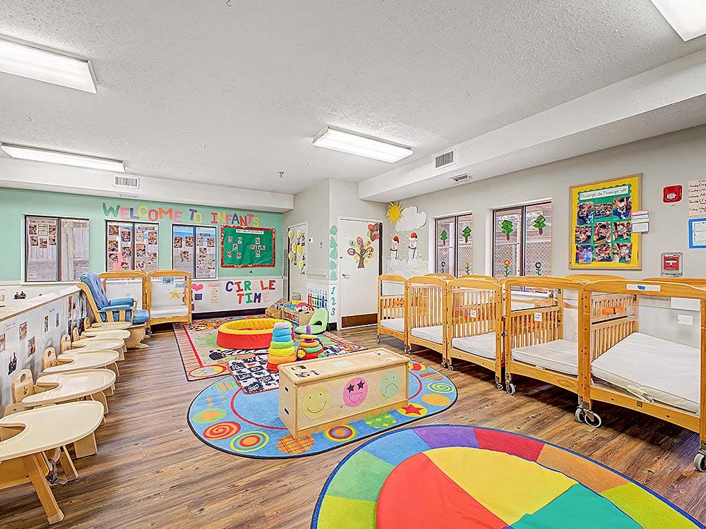 An infant daycare room with several cribs, high chairs, and colorful rugs on the floor