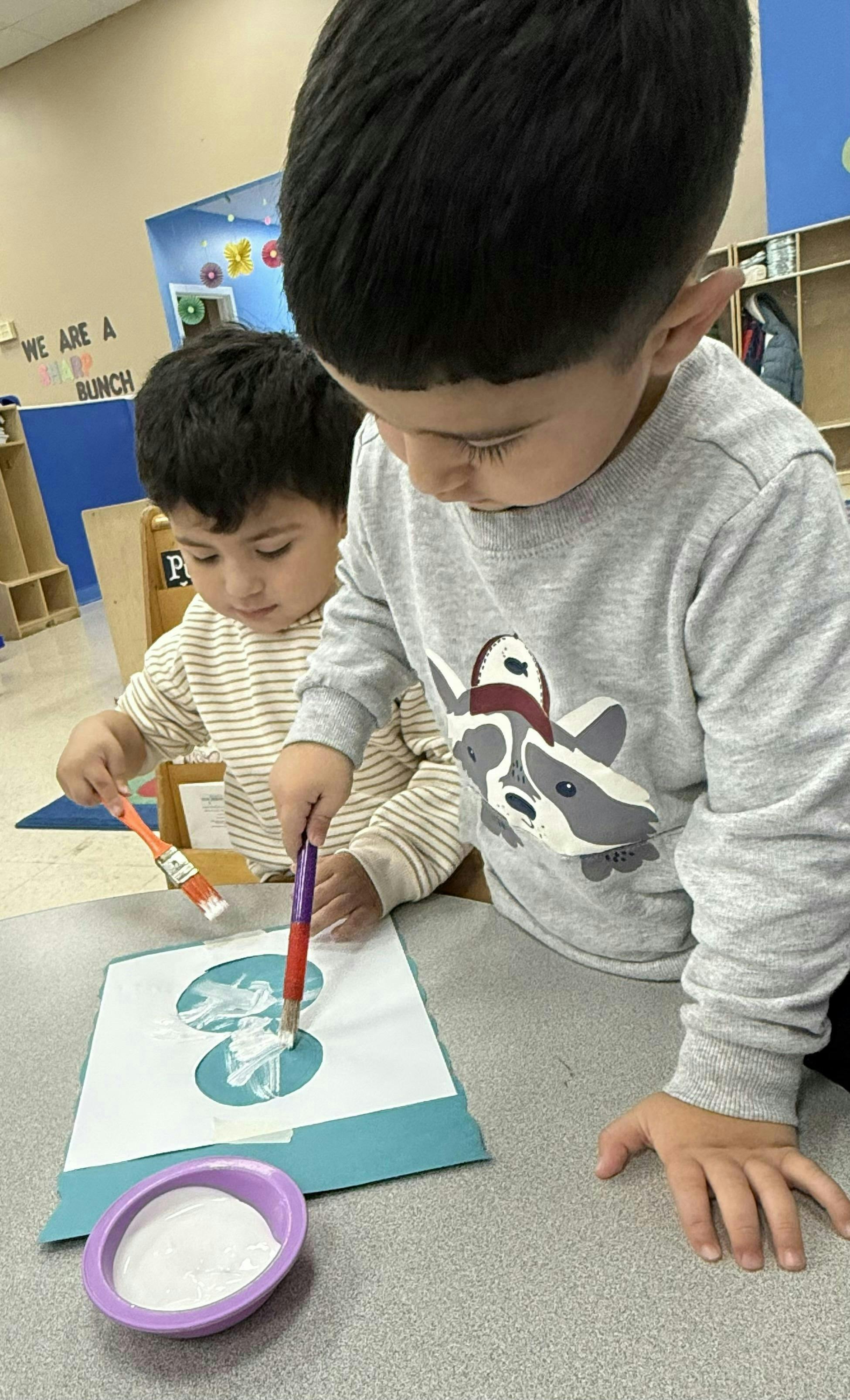 Two children painting with white paint and brushes at a table in a classroom