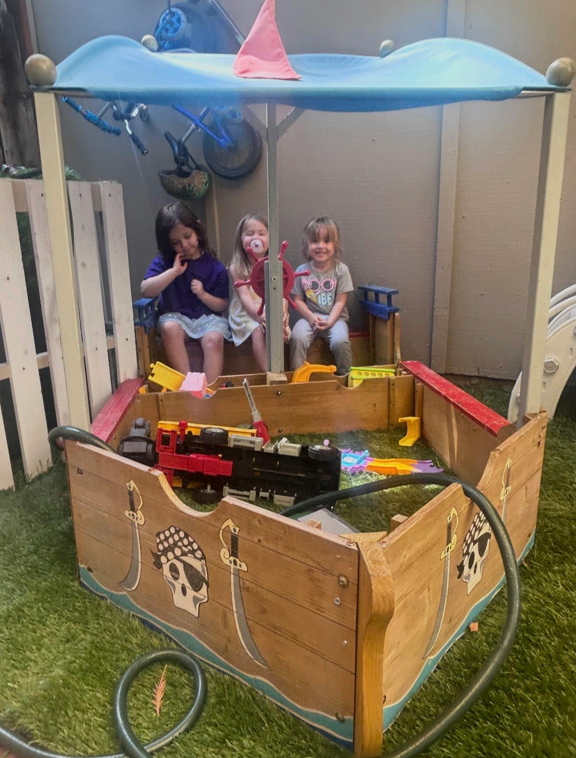 Three children sitting inside a wooden pirate ship play structure