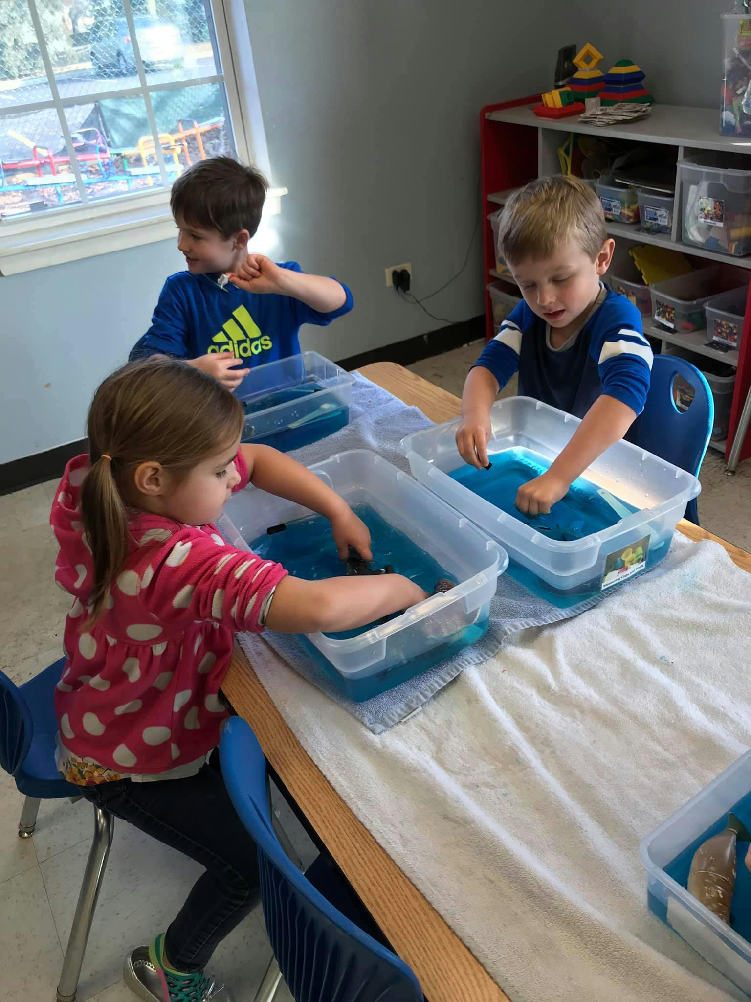 Three children playing with blue liquid in plastic bins at a table