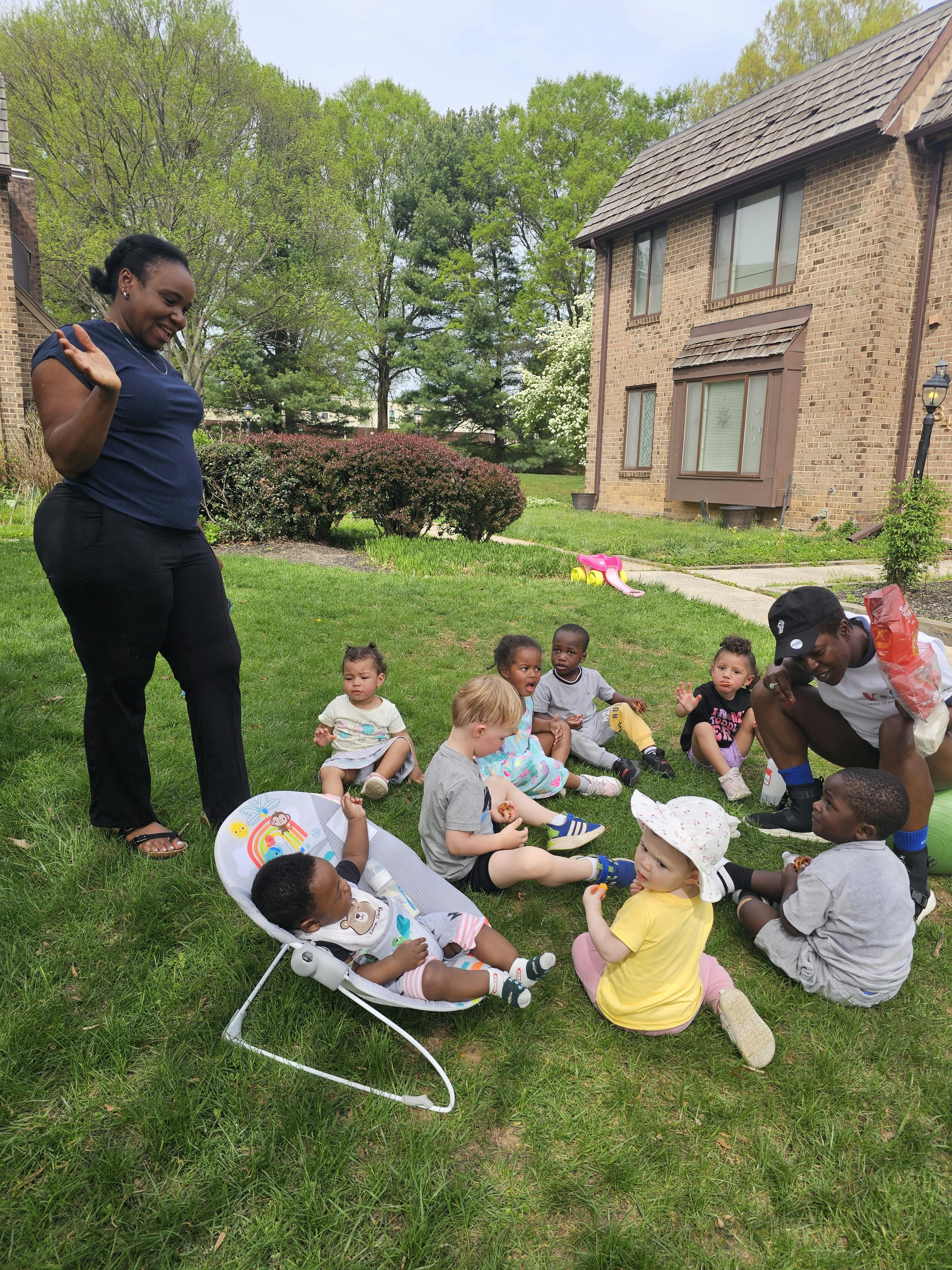 Adults and children gathered on a grassy lawn with a building in the background