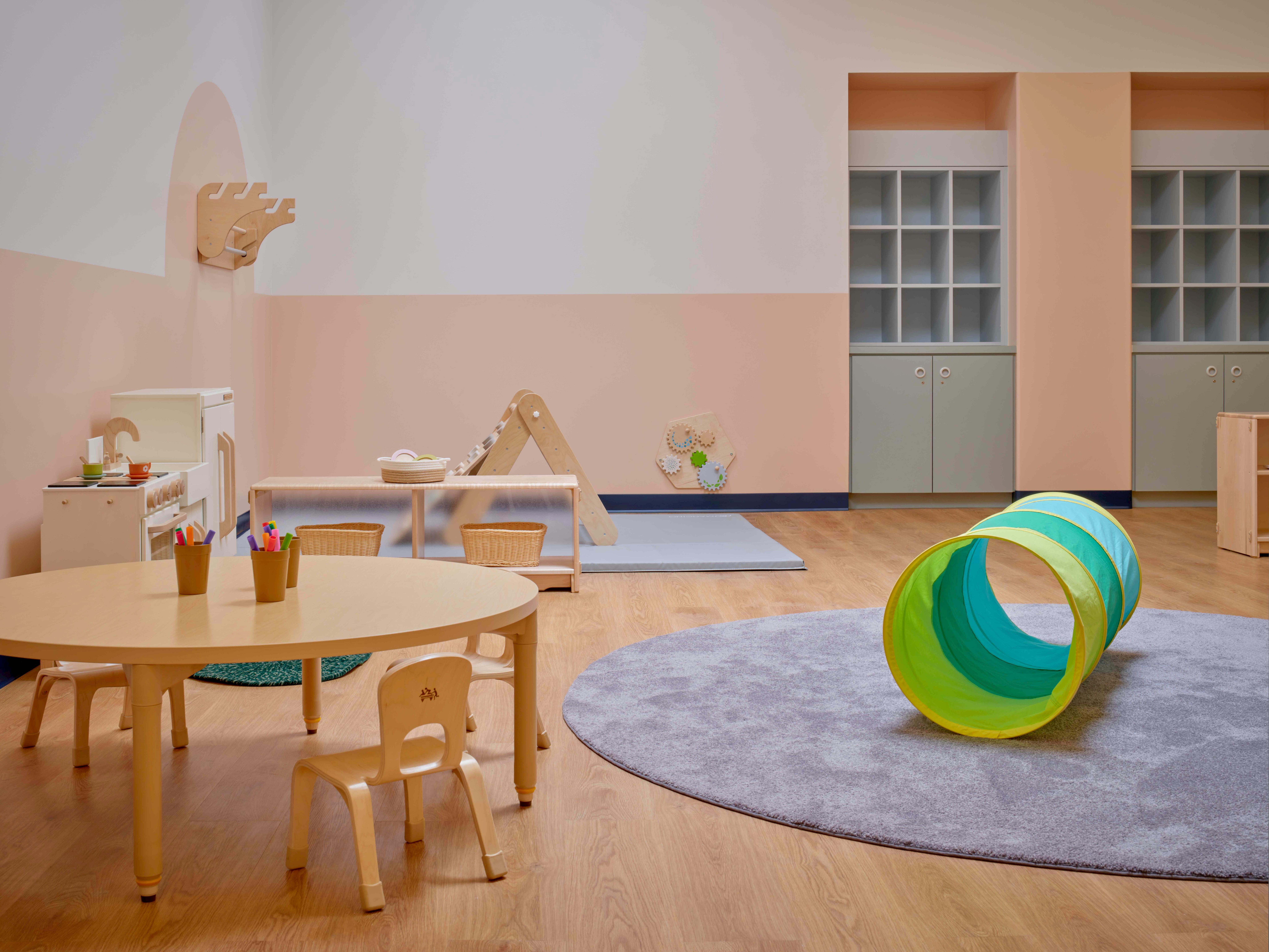 Playroom with small tables, chairs, and a colorful tunnel on a rug