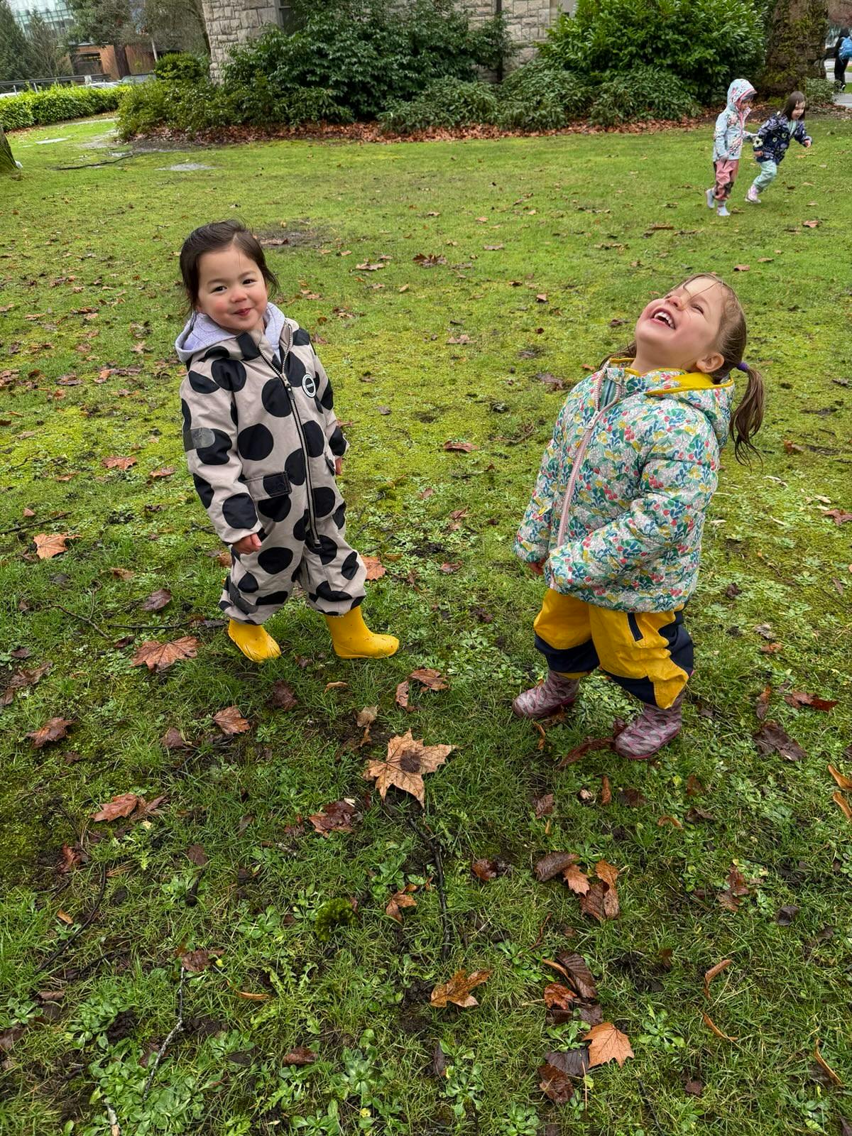 Two children in winter gear on a grassy field with other children in background