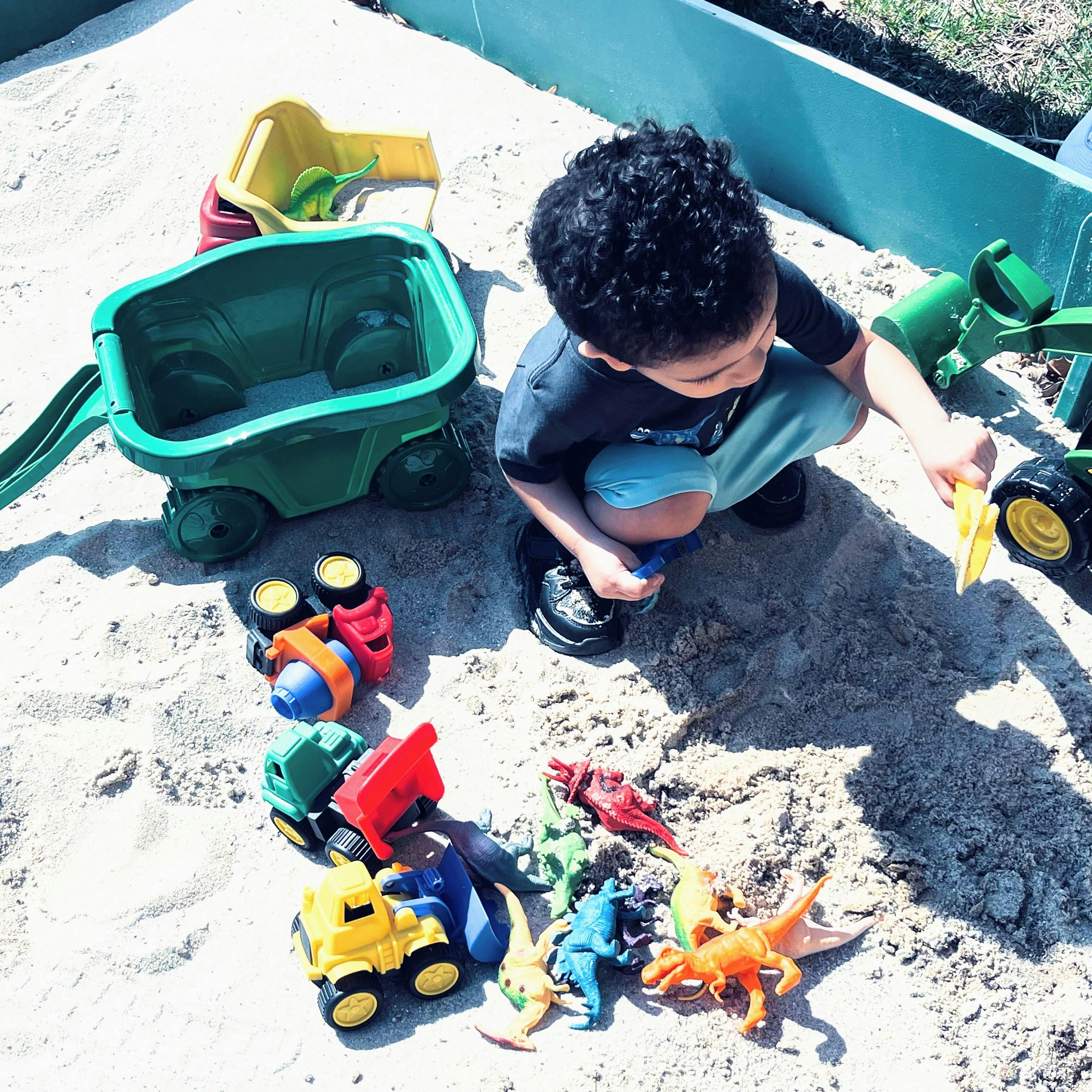 Child playing in a sandbox with toy trucks and dinosaur figures