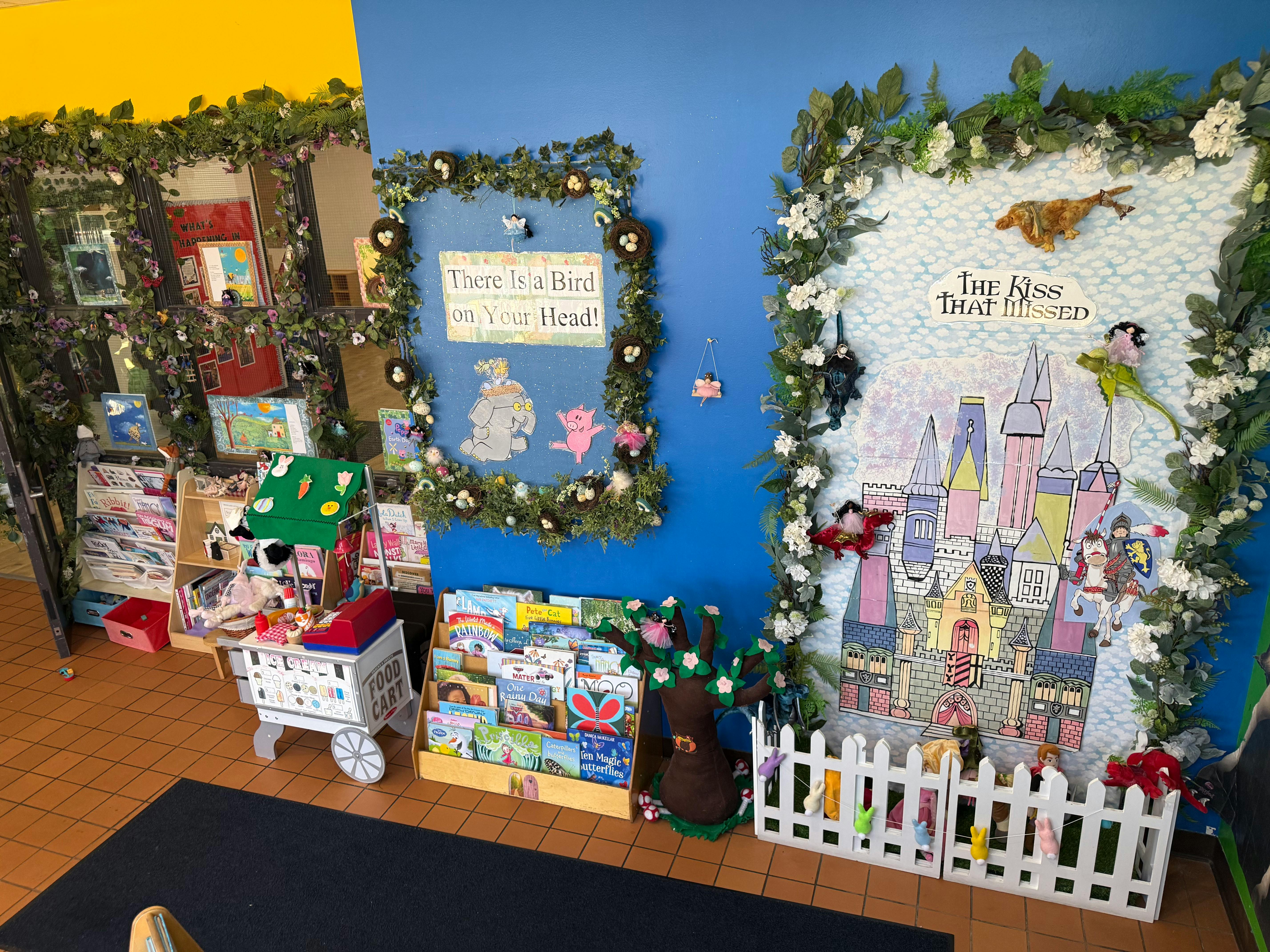 Children's corner with bookshelves, a play food cart, and fairy tale themed wall decor