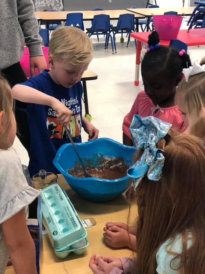 Child stirring batter in a blue bowl with other children watching