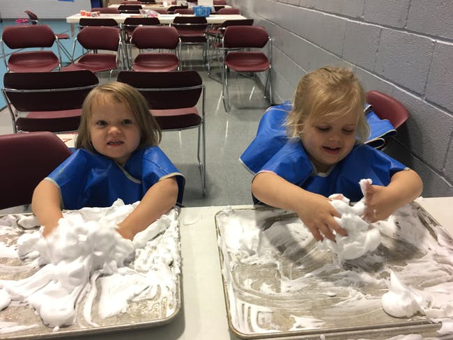Two children in blue smocks playing with shaving cream on trays