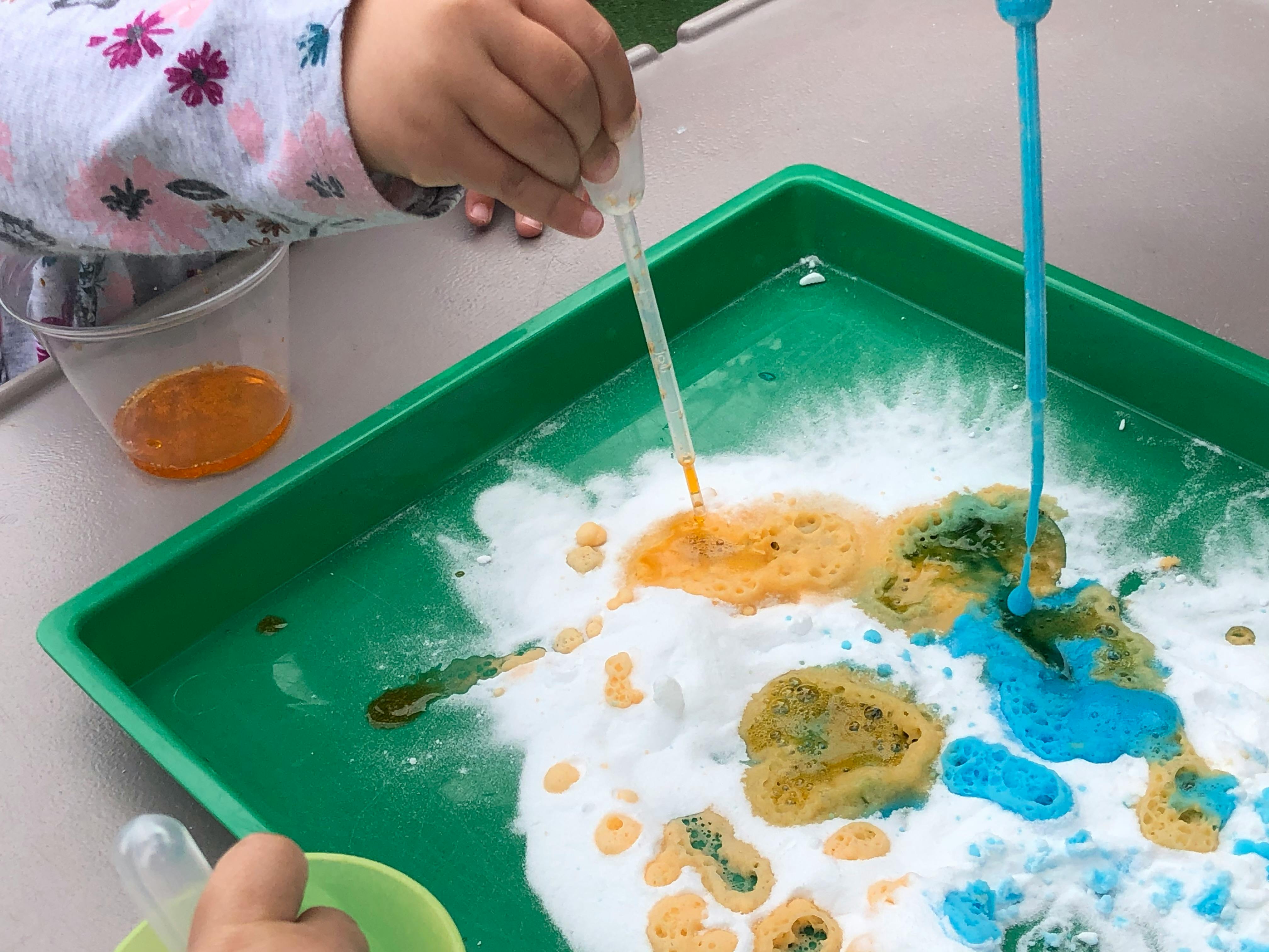 Child using a dropper to add liquid to a white powder reaction on a green tray